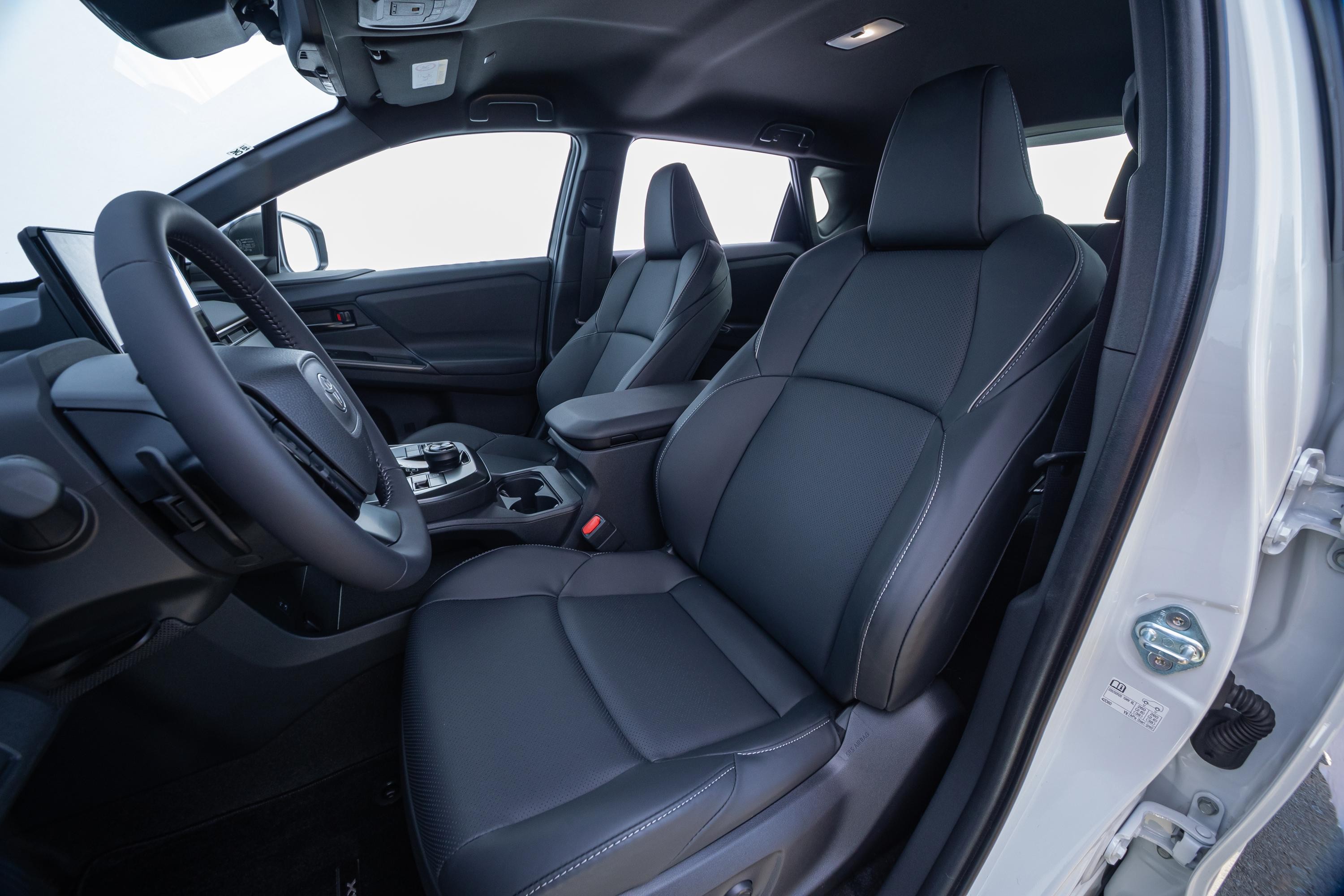 Interior de um carro moderno com bancos de couro escuro e costuras brancas, volante &agrave; esquerda, console central com porta-copos e tela digital no painel. A porta do passageiro est&aacute; aberta, revelando a estrutura interna do ve&iacute;culo.