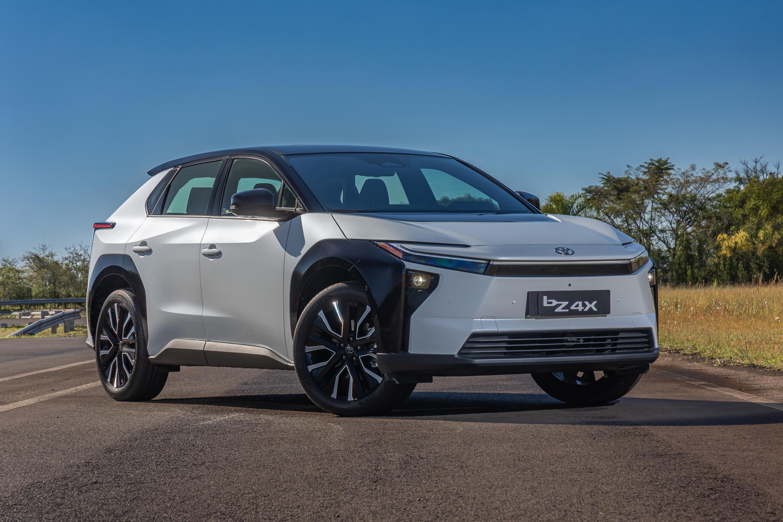 SUV el&eacute;trico Toyota bZ4X branco e preto, estacionado em uma estrada asfaltada, com vegeta&ccedil;&atilde;o e c&eacute;u azul ao fundo