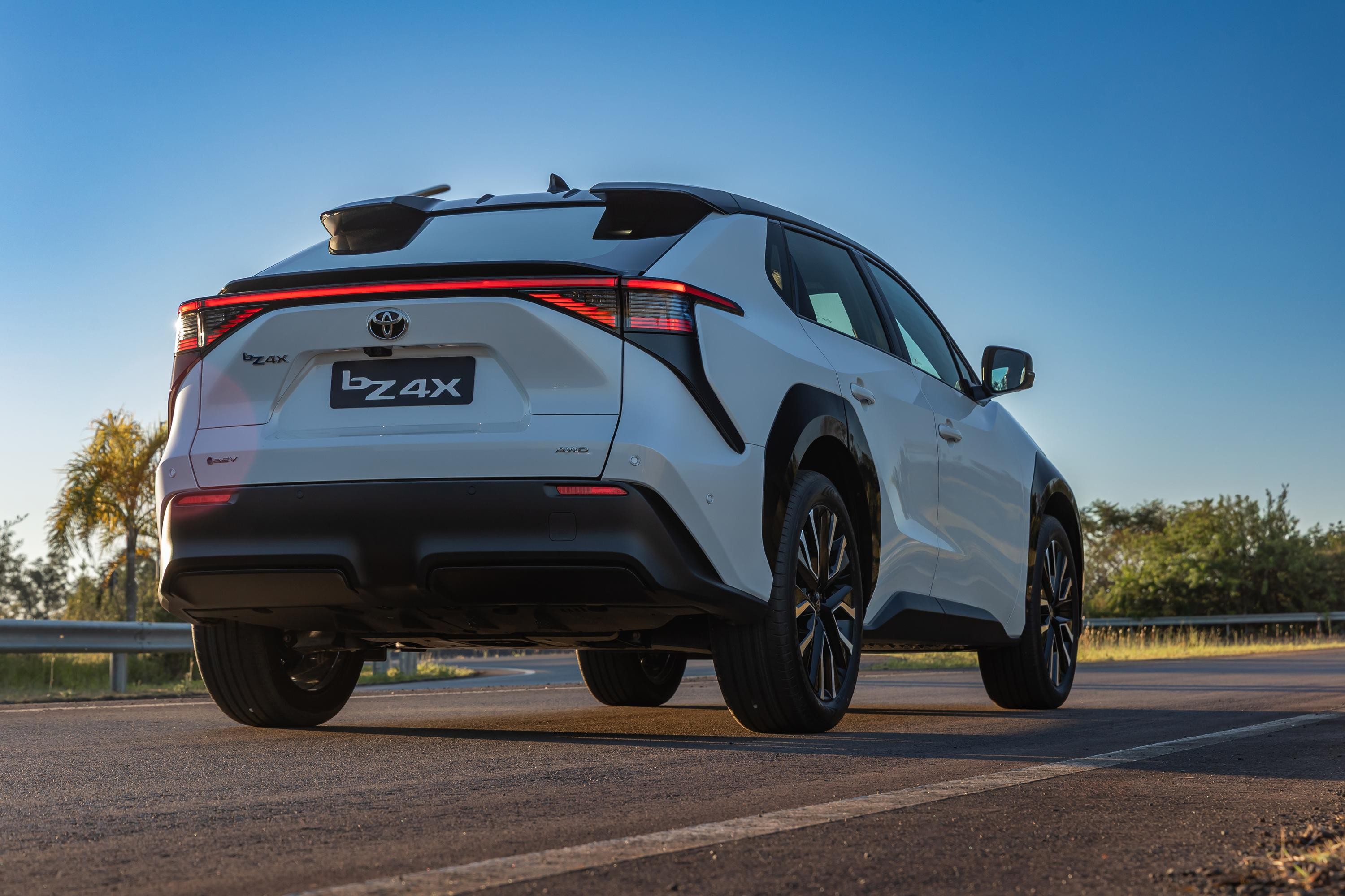 SUV Toyota bZ4X branco, visto por tr&aacute;s, parado em uma estrada asfaltada com faixa branca. O carro tem lanternas traseiras em LED vermelhas, para-choque preto e rodas escuras. Ao fundo, c&eacute;u azul claro e vegeta&ccedil;&atilde;o verde com uma palmeira &agrave; esquerda
