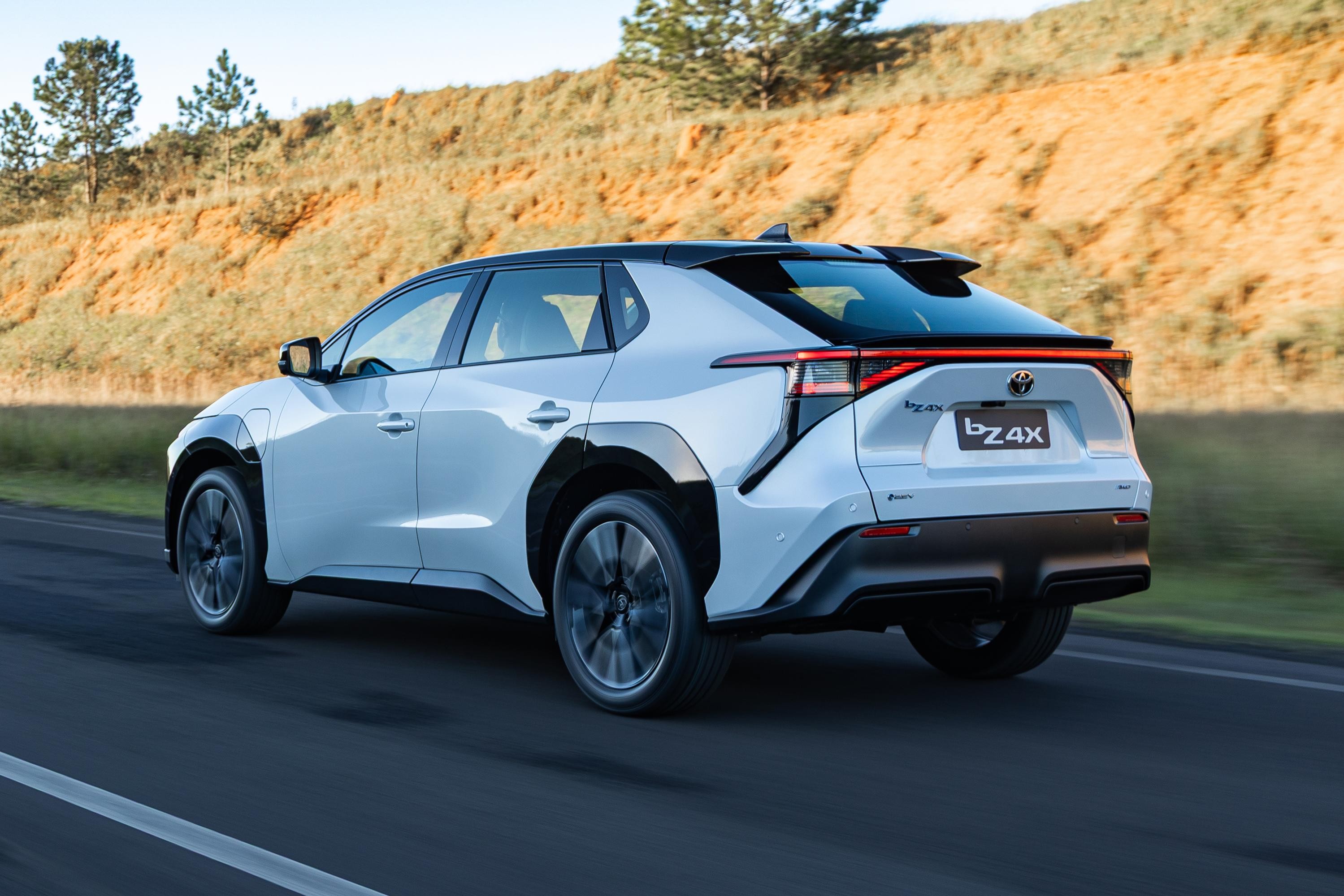 SUV el&eacute;trico Toyota bZ4X branco em movimento, visto de traseira lateral, com rodas escuras e teto preto, em estrada asfaltada com faixa branca. Ao fundo, um morro com vegeta&ccedil;&atilde;o seca e algumas &aacute;rvores verdes no topo