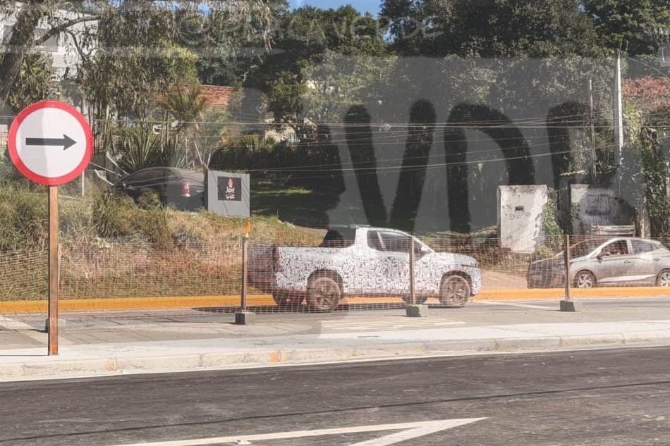 Sinal de tr&acirc;nsito circular branco e vermelho com seta preta apontando para a direita, em uma rua com asfalto novo e marca&ccedil;&otilde;es brancas. Ao fundo, uma picape camuflada e um carro cinza estacionados, com vegeta&ccedil;&atilde;o e casas ao redor sob c&eacute;u azul