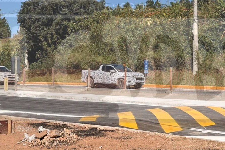 Duas picapes camufladas, uma branca e outra cinza, estacionadas em uma rua com faixa de pedestres amarela e branca, ladeada por vegeta&ccedil;&atilde;o densa e um poste de energia, sob c&eacute;u azul claro