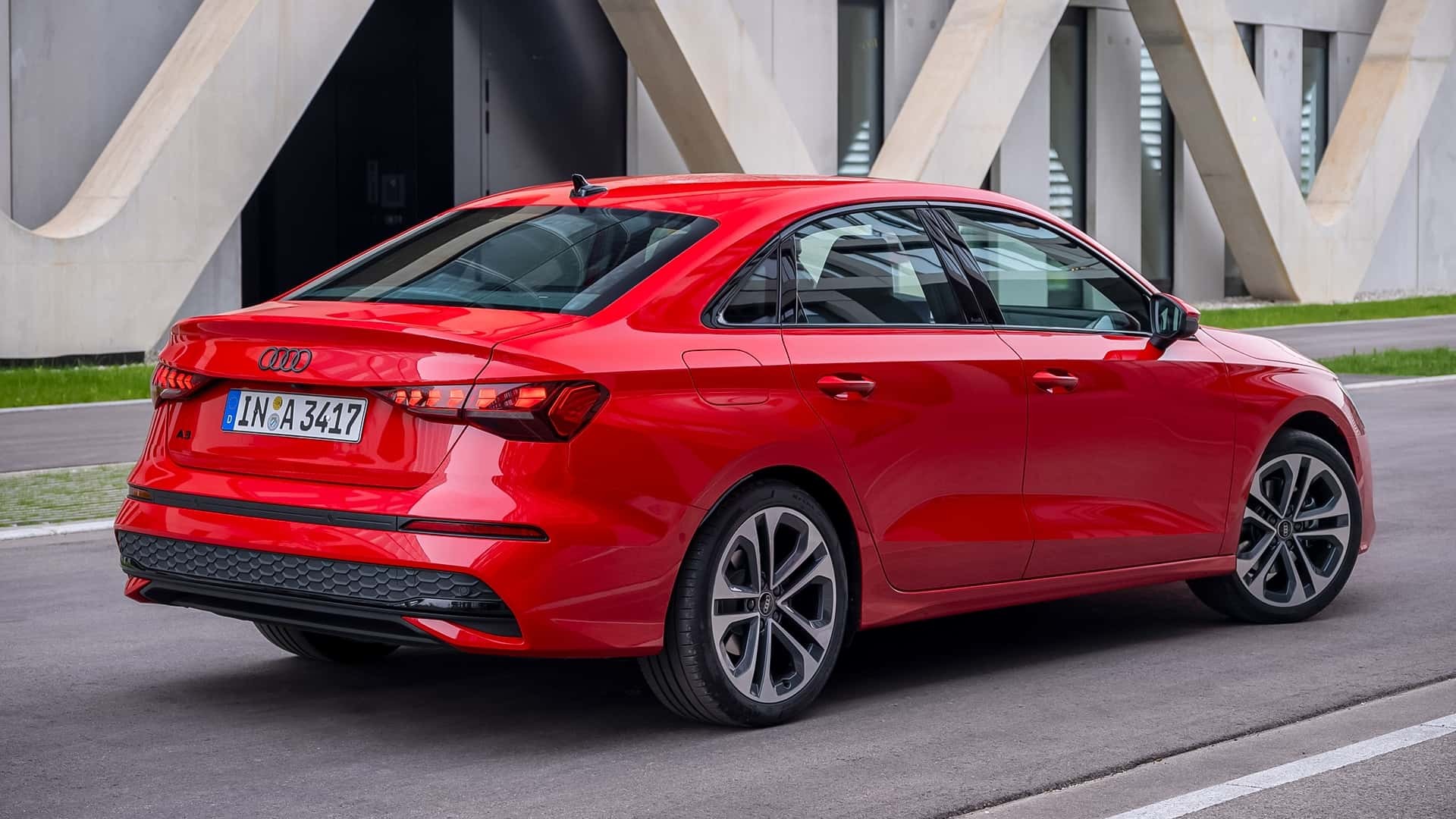 Carro Audi A3 Sedan vermelho estacionado em uma rua, visto de traseira e lateral, com um pr&eacute;dio moderno de concreto ao fundo