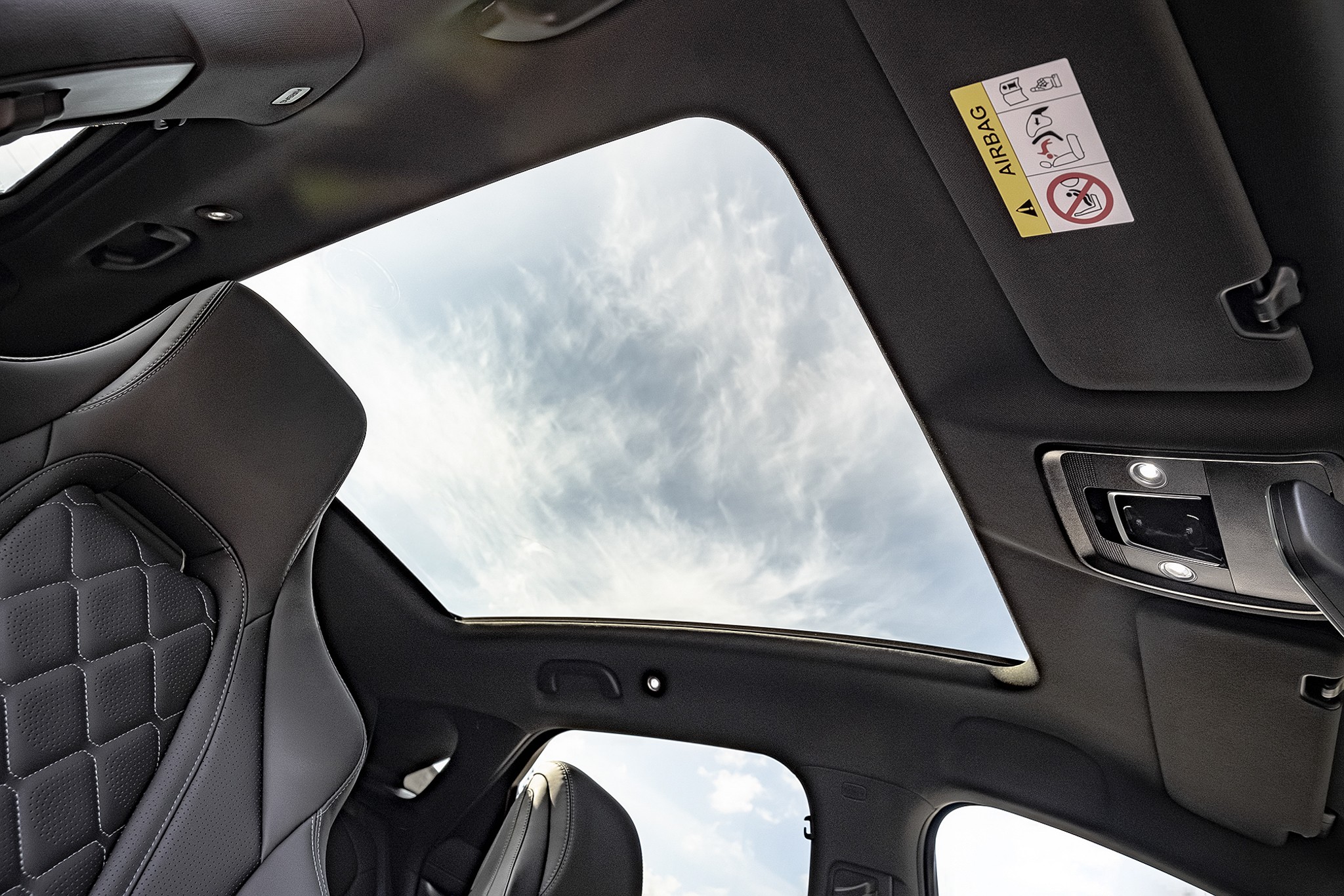 Interior de um carro com teto solar panor&acirc;mico, mostrando o c&eacute;u azul com nuvens esparsas. &Agrave; esquerda, parte do banco de couro preto com costura em losangos. &Agrave; direita, o console de luzes internas e o para-sol com adesivo de aviso do airbag