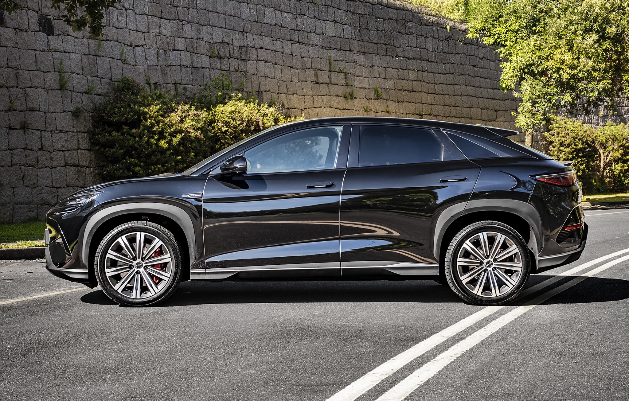 SUV preto brilhante estacionado de perfil em uma rua asfaltada, com uma parede de pedra e vegeta&ccedil;&atilde;o ao fundo. O carro tem rodas esportivas prateadas com detalhes vermelhos nos freios e acabamentos cinzas nas caixas de roda. Duas faixas brancas paralelas marcam o asfalto em primeiro plano