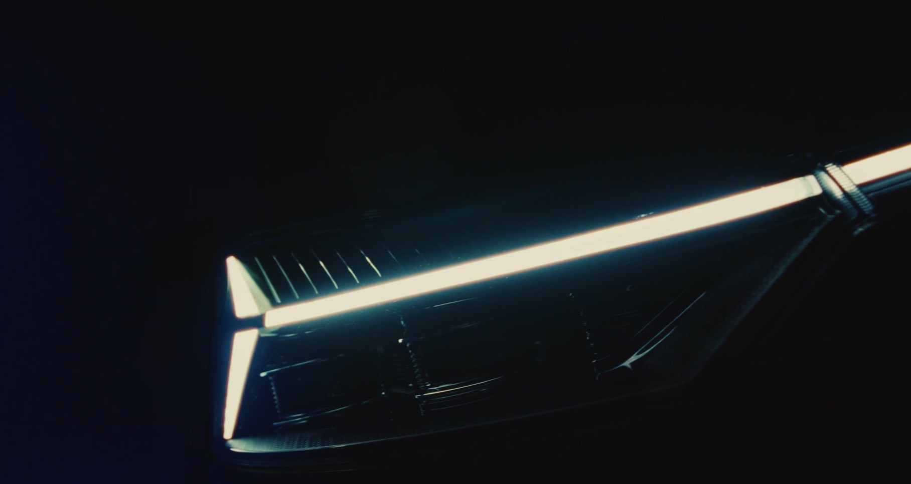 Detalhe de um farol de carro moderno com luzes LED brancas em formato de cruz e uma barra horizontal, em fundo escuro
