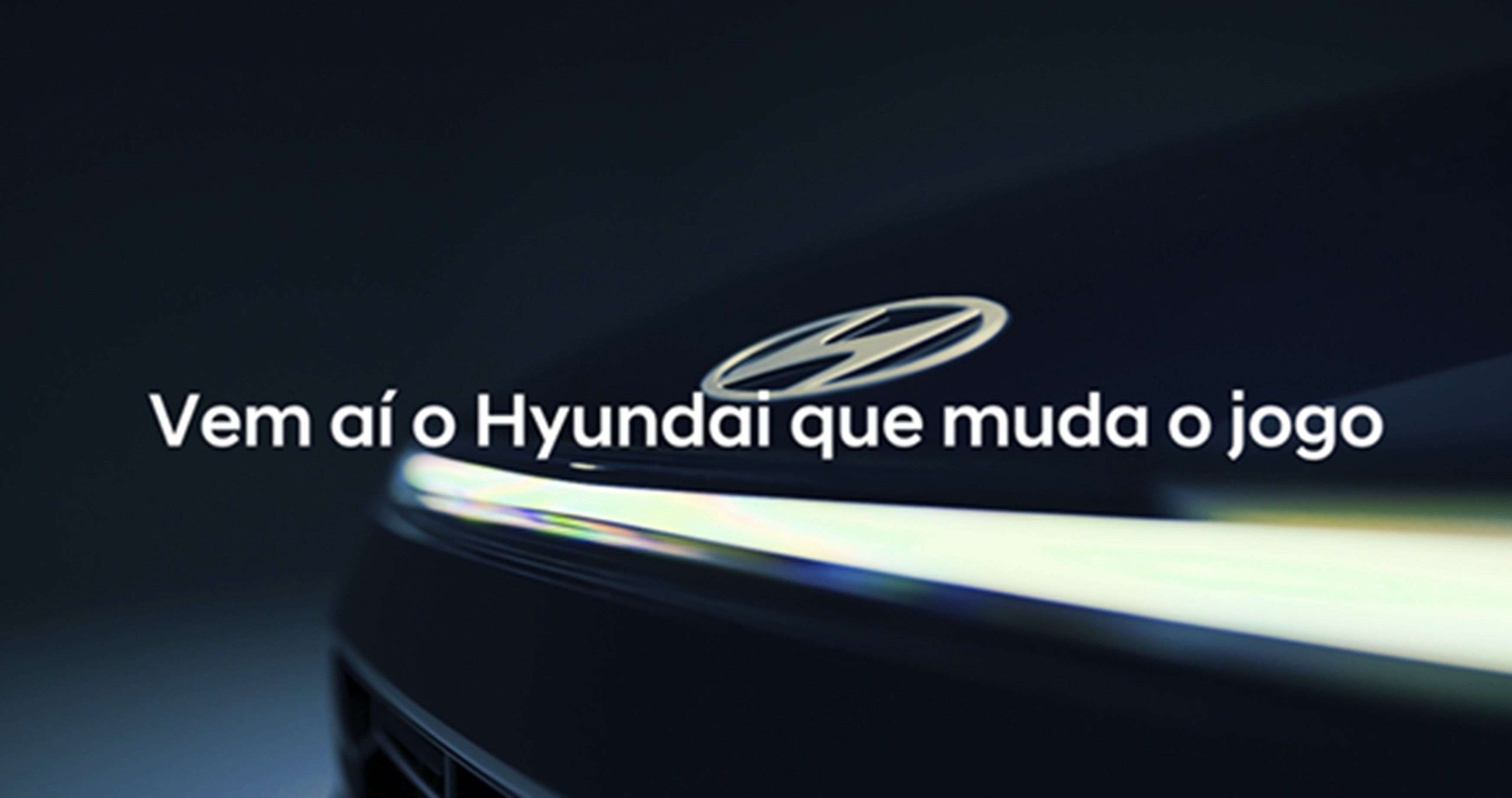 Parte frontal de um carro Hyundai escuro com o logotipo prateado e um farol de LED aceso, com o texto Vem a&iacute; o Hyundai que muda o jogo
