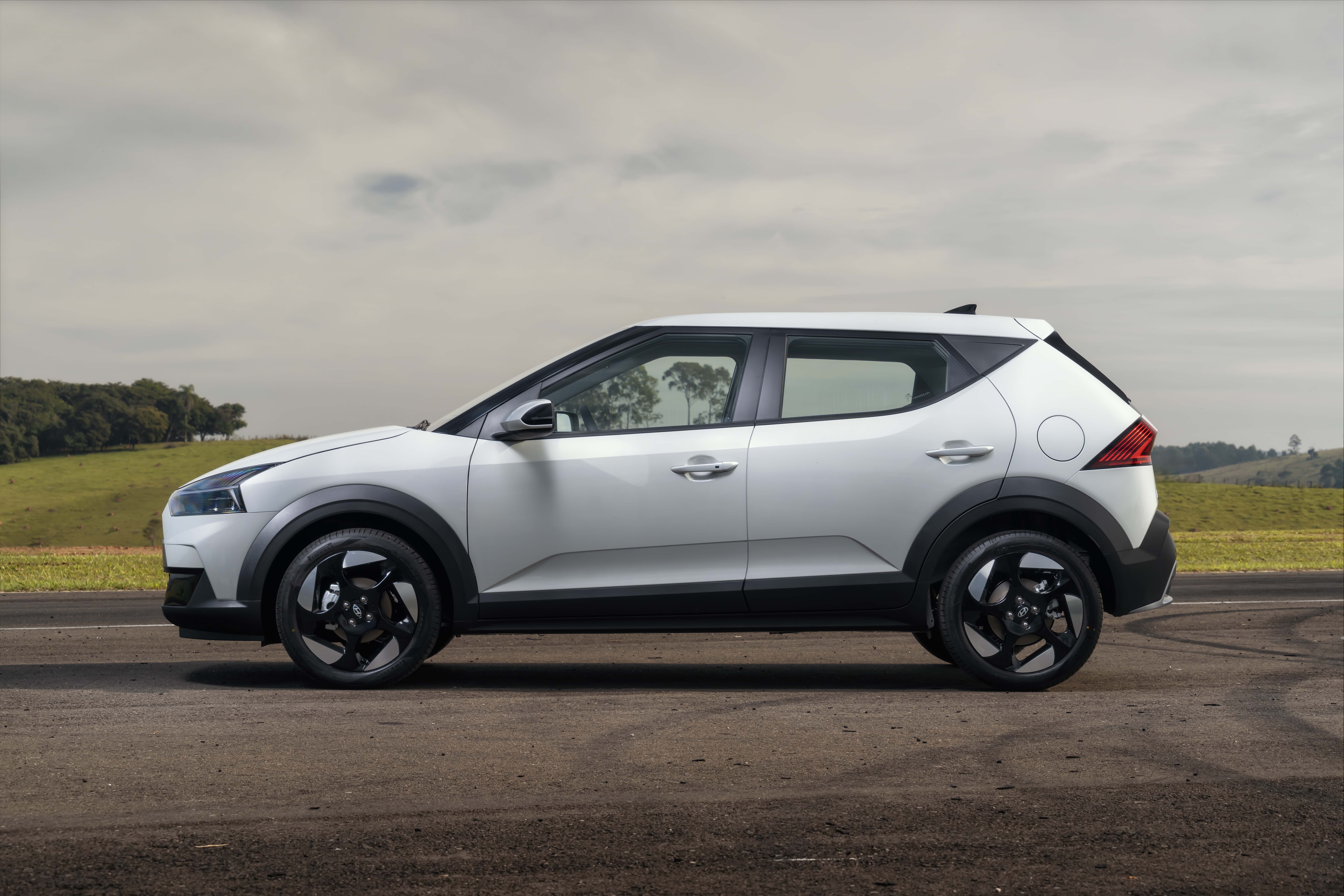 Carro SUV branco com detalhes pretos, rodas escuras e design moderno, estacionado em uma estrada de terra com paisagem rural ao fundo, sob c&eacute;u nublado