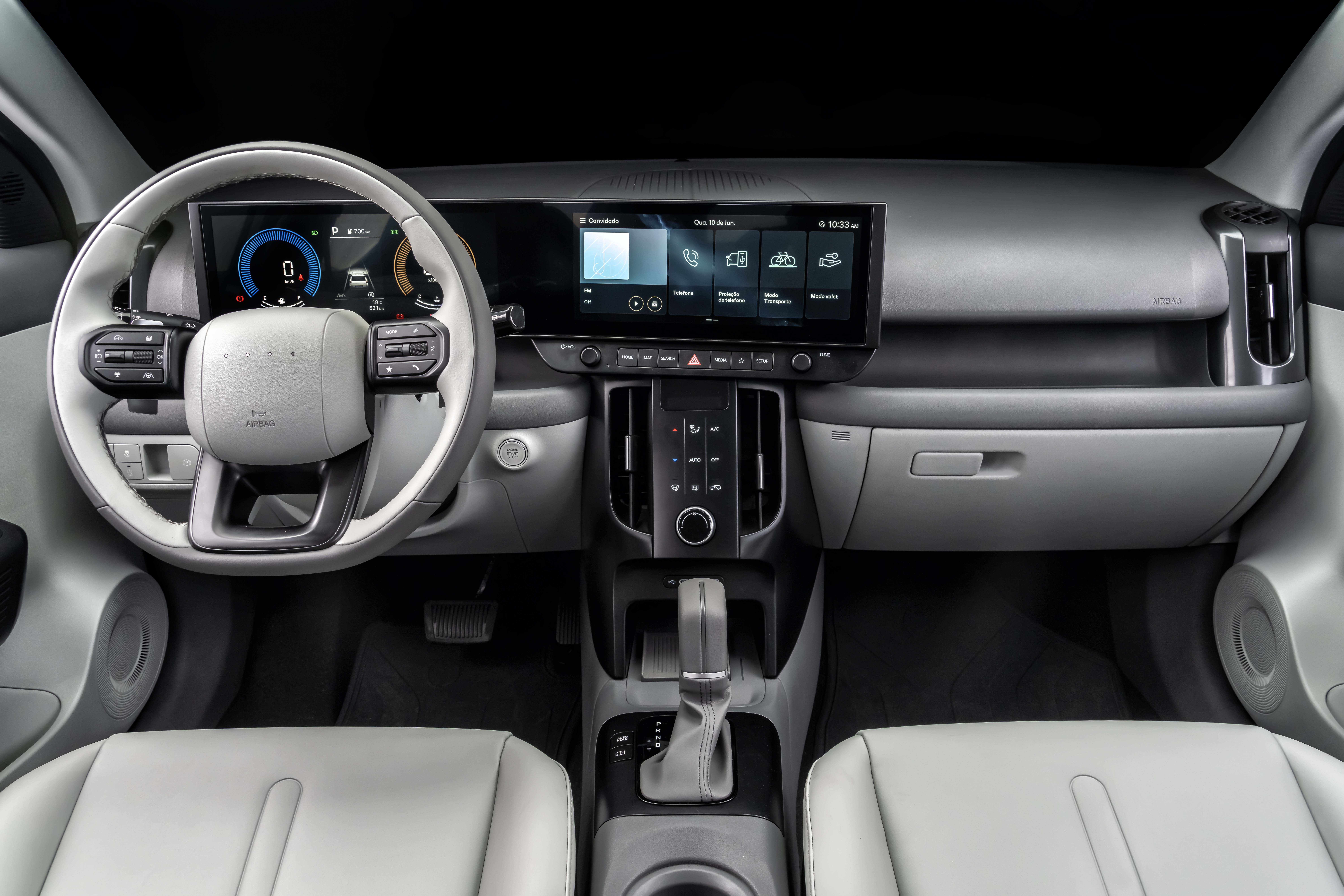 Interior de um carro moderno com volante branco e cinza, painel digital com veloc&iacute;metro e tela multim&iacute;dia exibindo &iacute;cones de aplicativos. O console central tem bot&otilde;es de controle e c&acirc;mbio autom&aacute;tico, com bancos de couro cinza claro vis&iacute;veis em primeiro plano