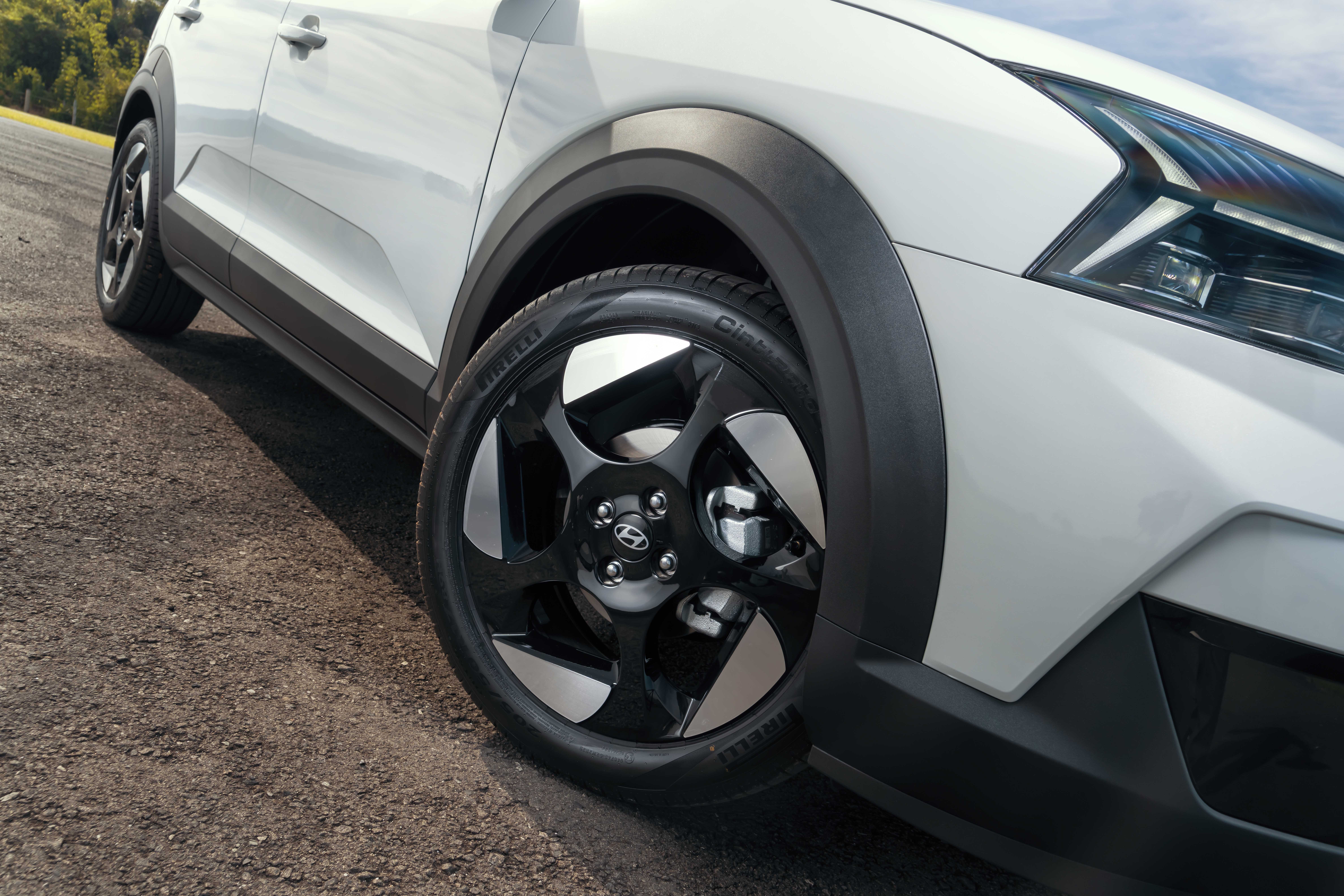 Lateral dianteira de um carro branco com detalhes pretos, mostrando a roda com aro escuro e detalhes prateados, pneu Pirelli e farol angular aceso, em uma estrada de terra