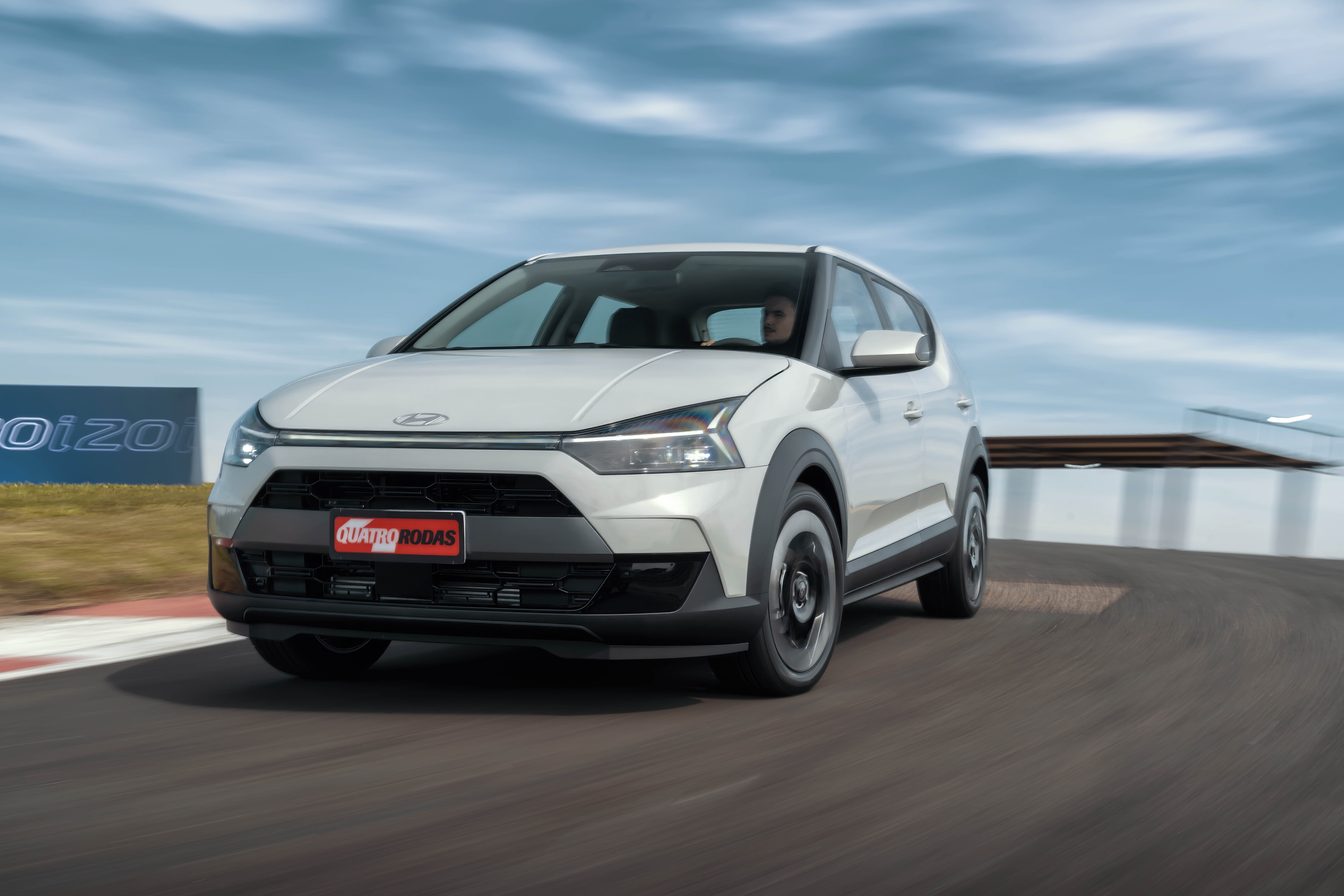 Carro branco Hyundai i20 em movimento numa pista de corrida, com um motorista vis&iacute;vel. O ve&iacute;culo tem detalhes em preto fosco e a placa Quatro Rodas na frente. O c&eacute;u azul com nuvens e uma placa azul com i20 ao fundo complementam a cena