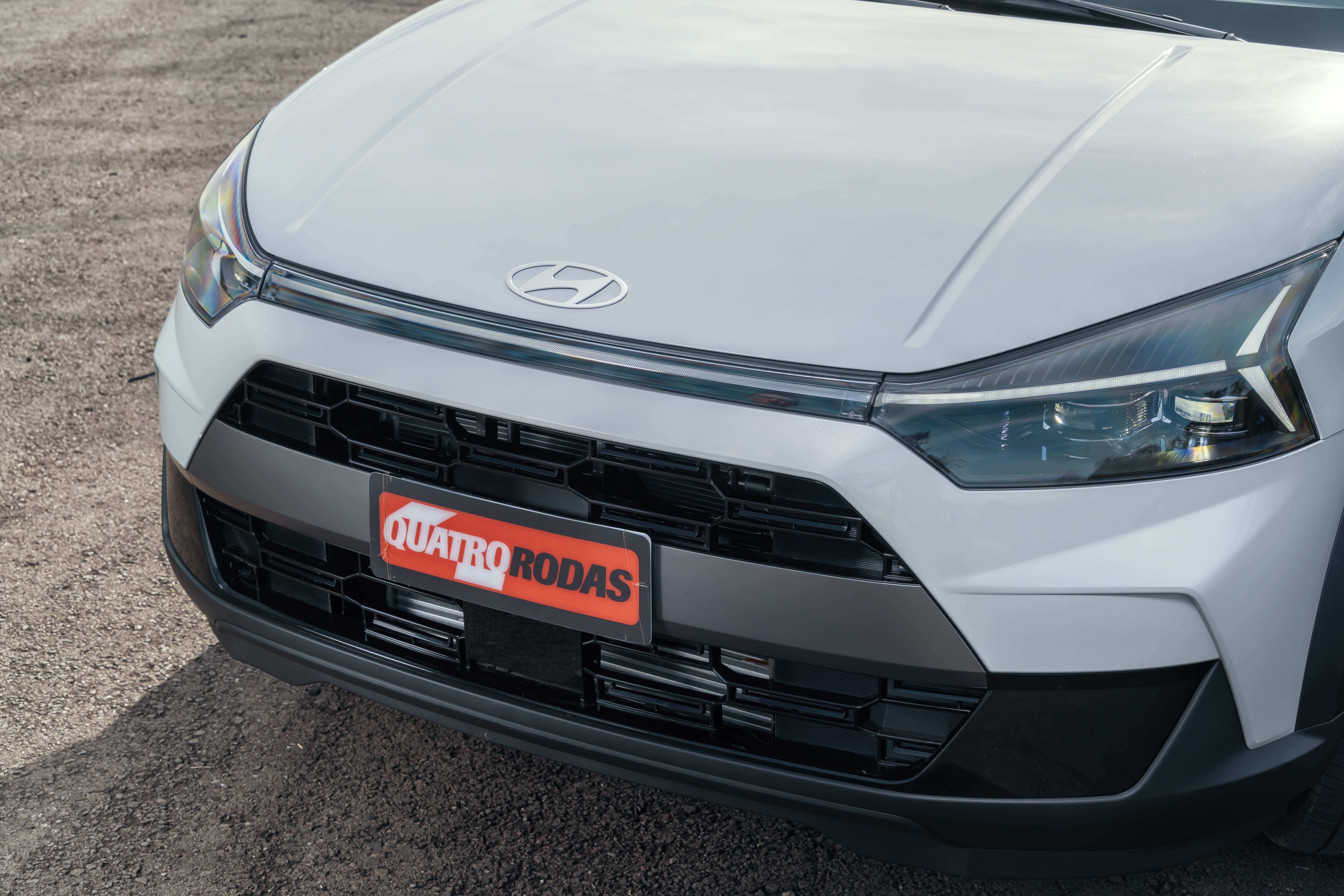 Parte frontal de um carro branco com o emblema da Hyundai no cap&ocirc;. A grade preta tem um padr&atilde;o hexagonal e uma placa vermelha com QUATRO RODAS em branco est&aacute; centralizada. Far&oacute;is alongados e modernos completam o design. O ve&iacute;culo est&aacute; sobre um piso de terra batida