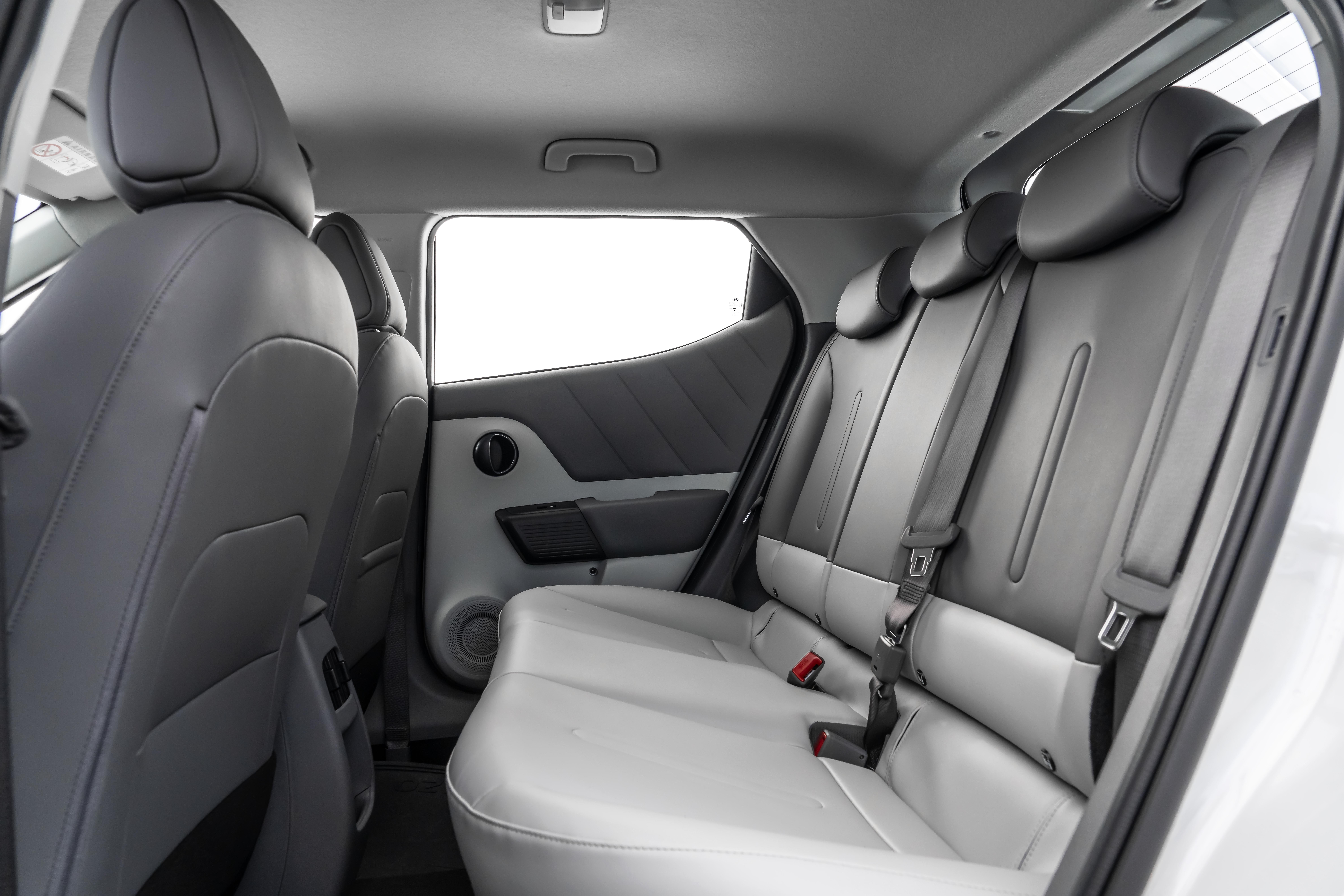 Interior de um carro com bancos traseiros cinza-escuro e cinza-claro, com tr&ecirc;s encostos de cabe&ccedil;a e cintos de seguran&ccedil;a. A porta traseira tem um painel cinza com detalhes em preto e uma janela retangular com fundo branco. O teto &eacute; cinza claro com uma al&ccedil;a de apoio.
