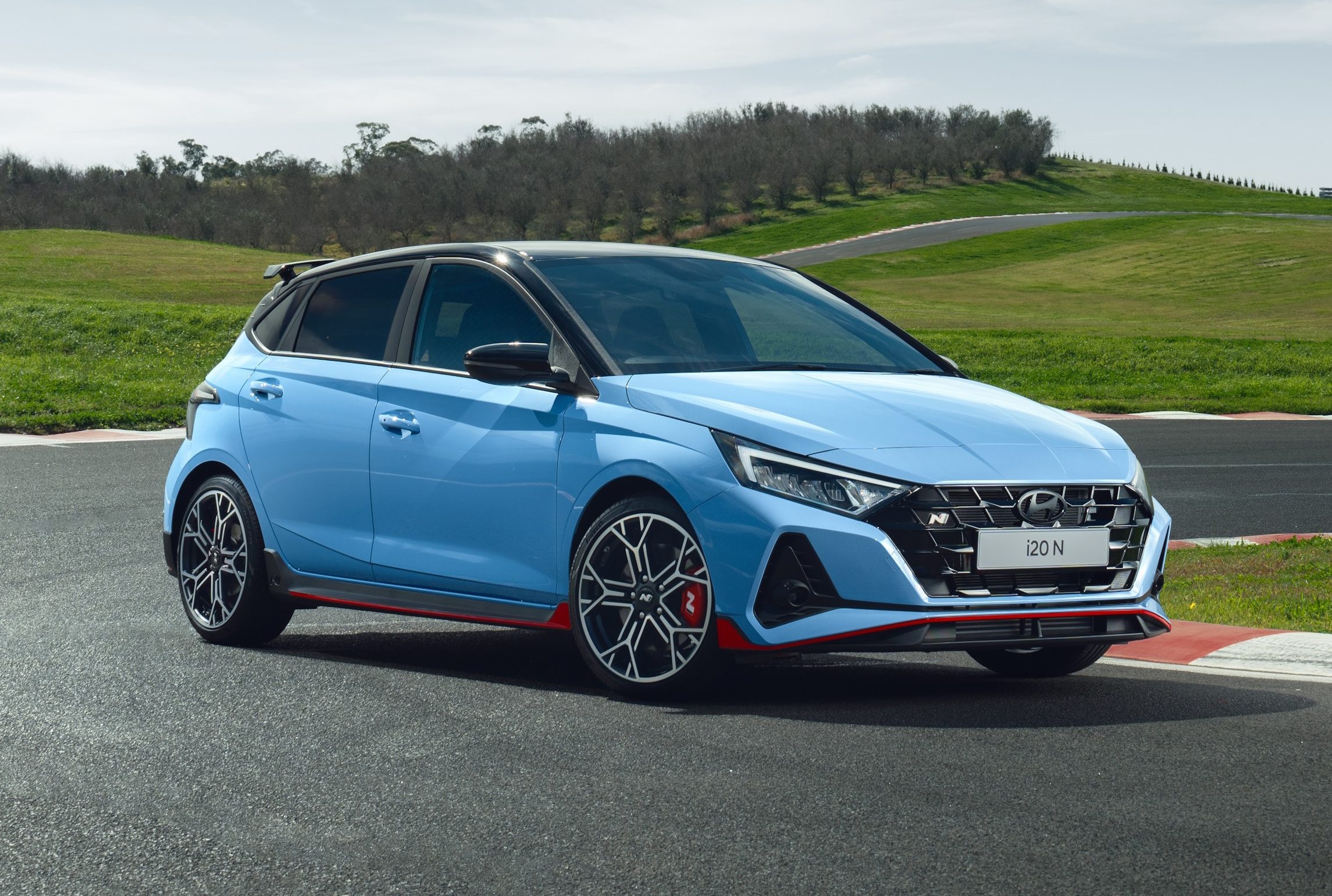 Hatchback azul claro com teto preto e detalhes vermelhos, parado em uma pista de corrida com grama verde e c&eacute;u azul ao fundo