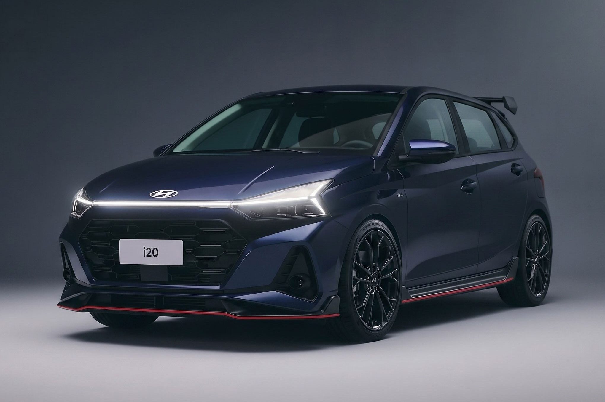 Hatchback Hyundai i20 azul-escuro com detalhes esportivos, como rodas pretas, saias laterais e spoiler traseiro, em um est&uacute;dio com fundo cinza-escuro. A placa frontal exibe i20