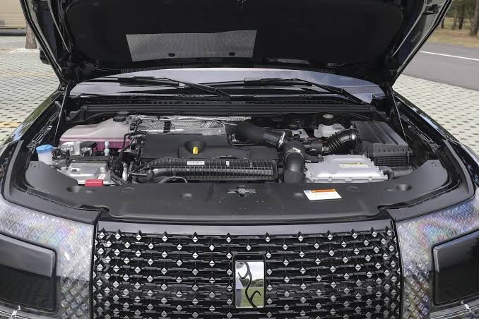 Motor de um carro preto com o cap&ocirc; aberto, revelando componentes como o reservat&oacute;rio de fluido rosa, bateria e mangueiras. A grade frontal possui um padr&atilde;o de losangos e um emblema central prateado. O ve&iacute;culo est&aacute; estacionado em um piso de blocos cinzas, com uma estrada e &aacute;rvores ao fundo