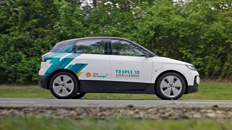 Carro el&eacute;trico branco com detalhes azuis e verdes, logotipos Shell Recharge e Triple 10 Challenge, parado em uma estrada asfaltada com vegeta&ccedil;&atilde;o ao fundo