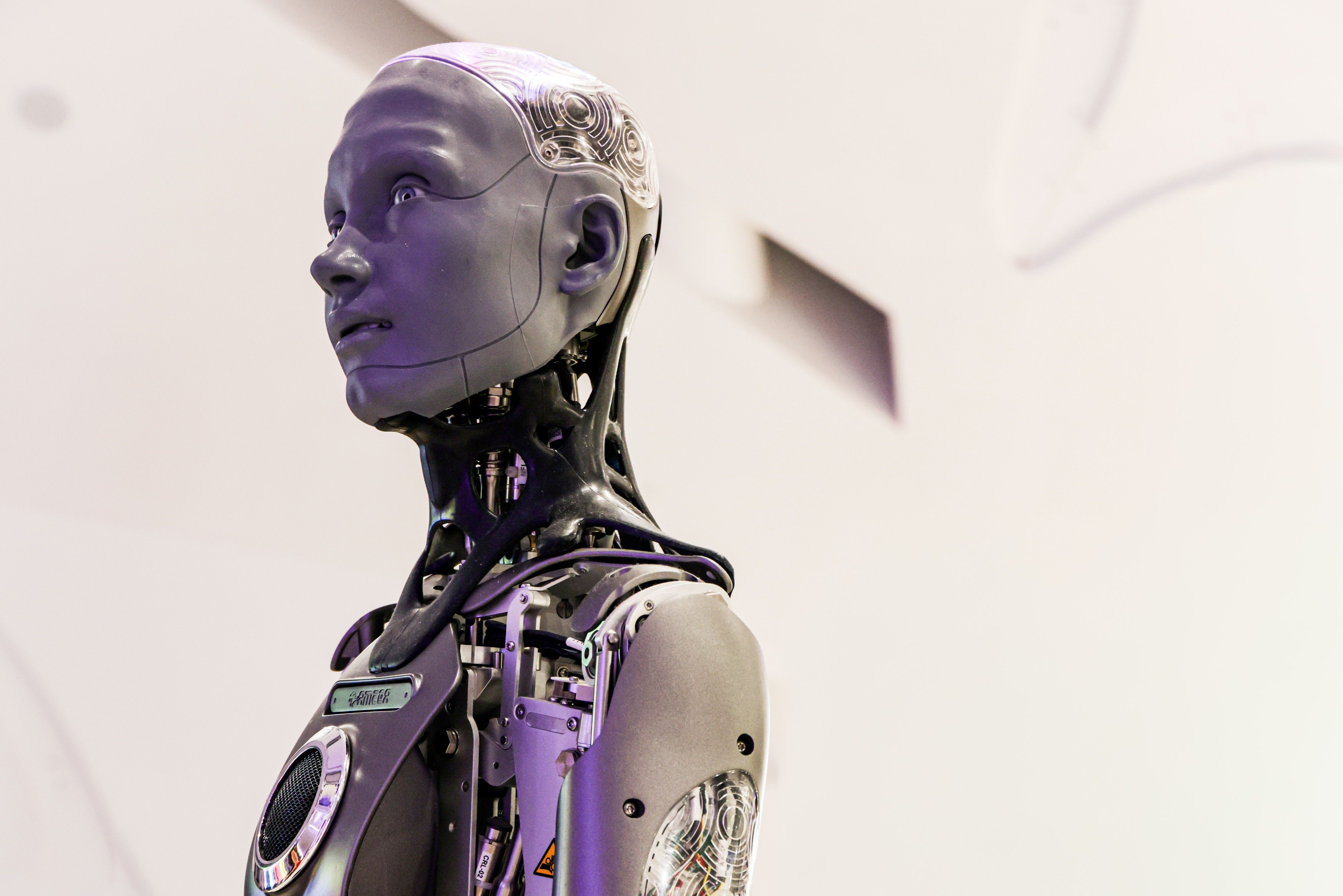 Rob&ocirc; humanoide prateado e preto, com detalhes de circuitos vis&iacute;veis, olhando para cima e para a direita, em um ambiente claro com cadeiras brancas ao fundo