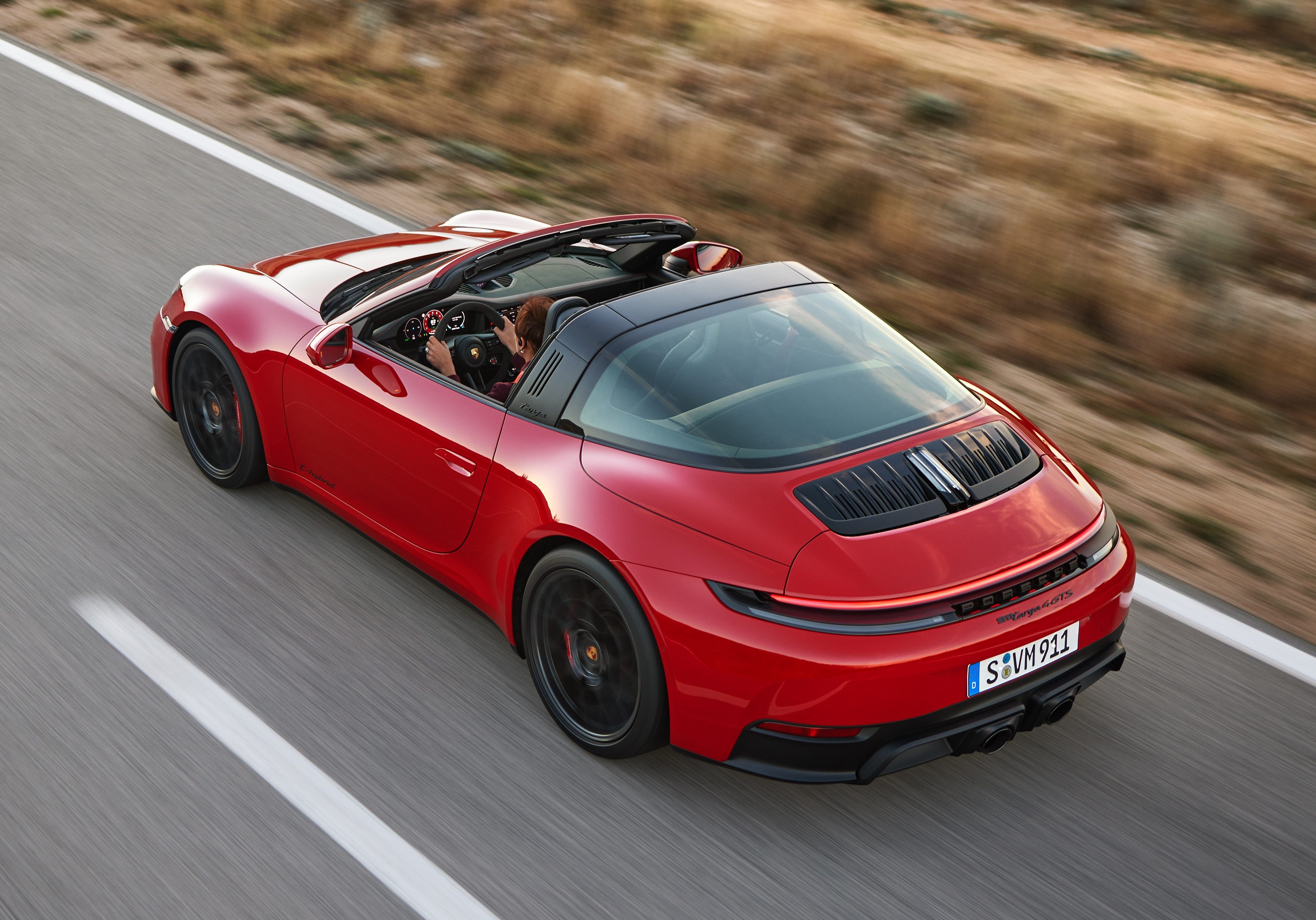 Carro esportivo vermelho, modelo Porsche 911 Targa, com teto convers&iacute;vel preto, em movimento r&aacute;pido numa estrada asfaltada com faixas brancas, ladeada por vegeta&ccedil;&atilde;o seca e borrada pelo movimento