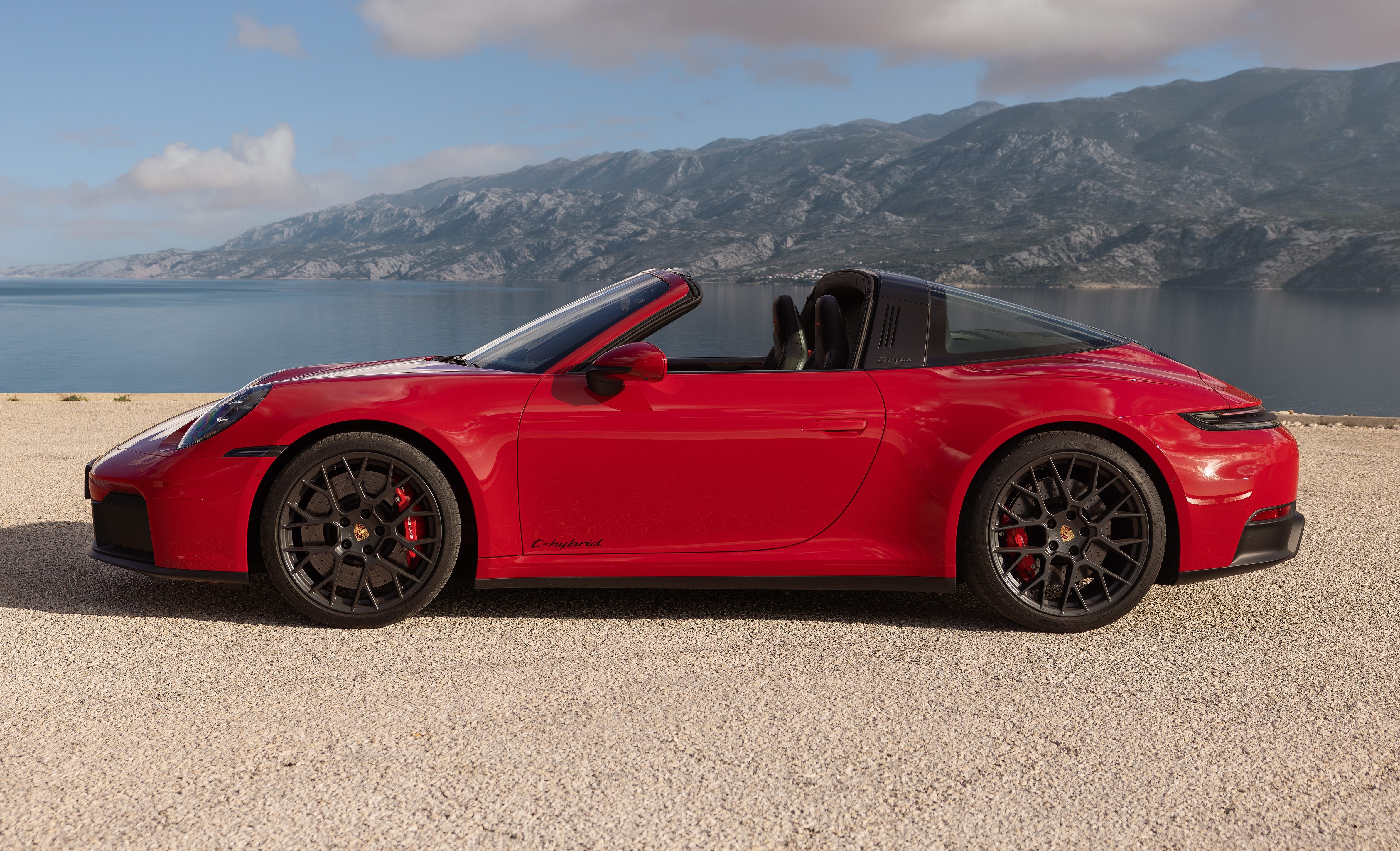 Carro esportivo vermelho, um Porsche 911 Targa, estacionado em uma estrada de terra com o teto retr&aacute;til aberto. Ao fundo, um lago azul e montanhas sob um c&eacute;u claro com nuvens brancas
