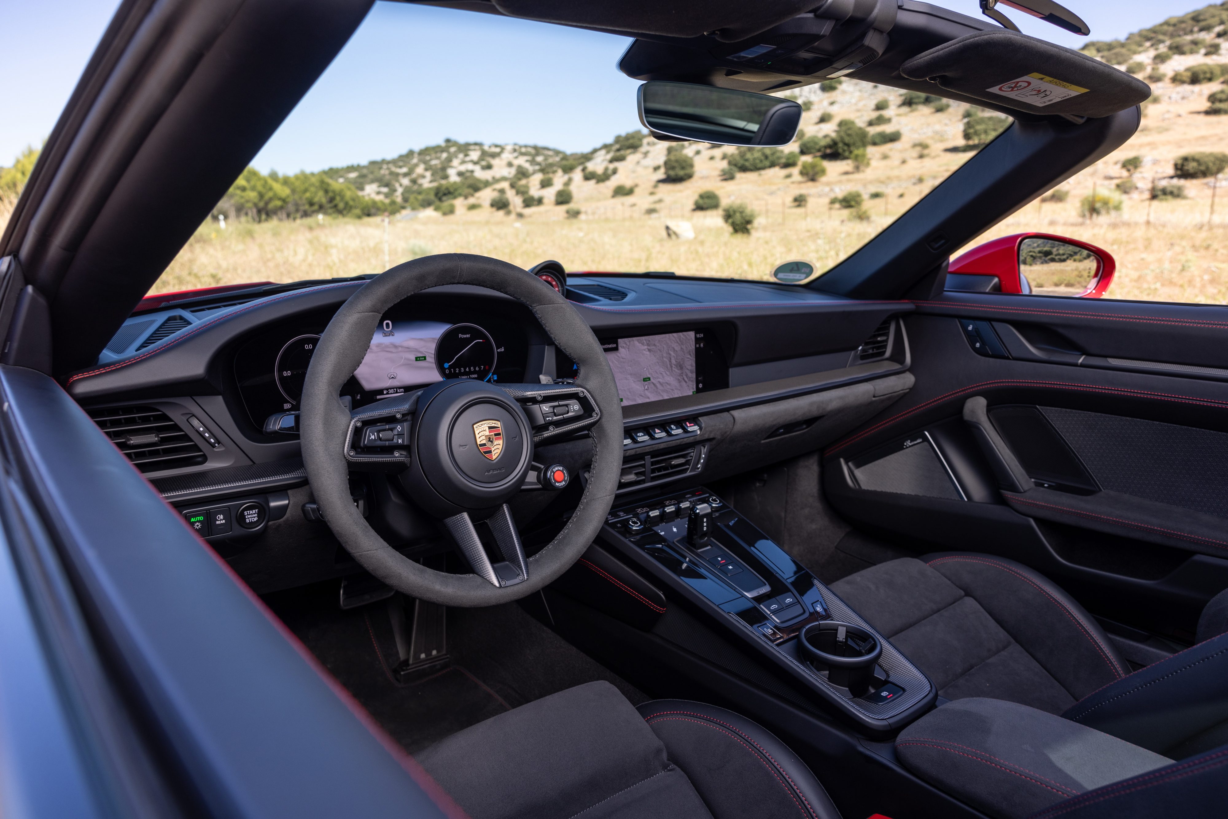 Interior de um carro esportivo convers&iacute;vel vermelho, com o volante e painel escuros em primeiro plano. O volante tem o emblema da Porsche. Ao fundo, uma paisagem montanhosa &aacute;rida sob c&eacute;u azul claro