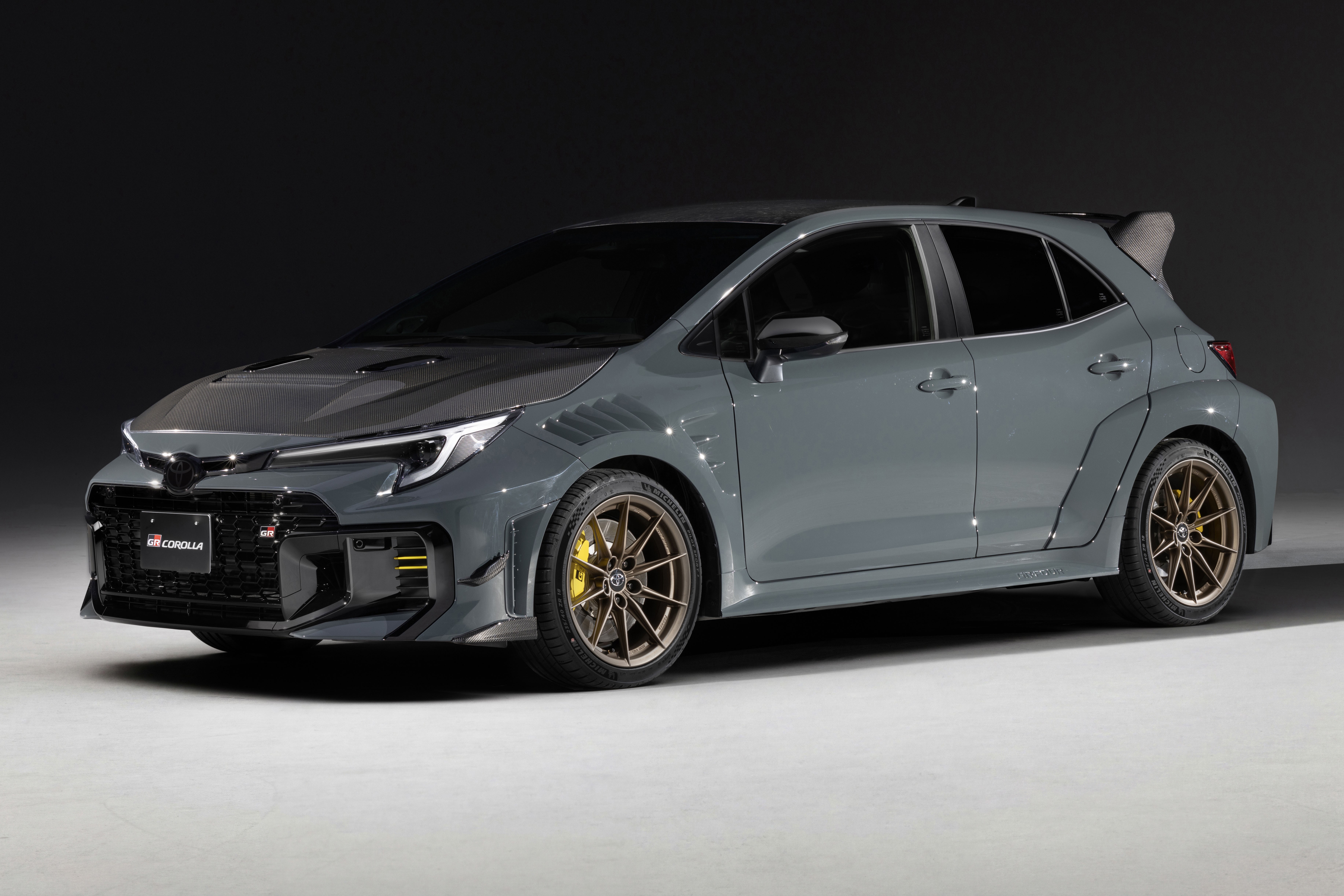 Carro esportivo cinza-chumbo, modelo hatchback, com cap&ocirc; escuro e rodas douradas, parado em est&uacute;dio com fundo escuro