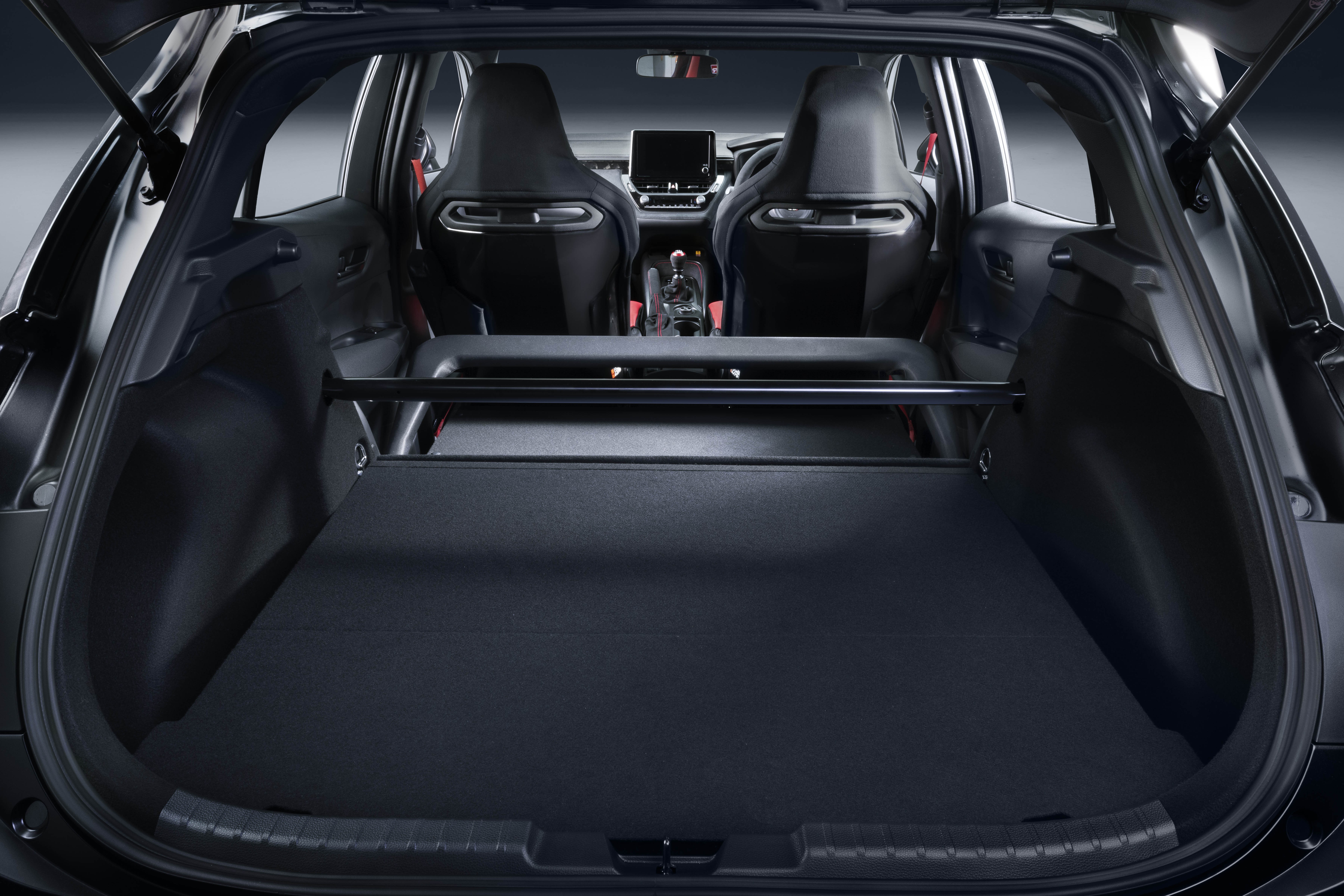 Interior de um carro esportivo preto, visto do porta-malas aberto. O porta-malas tem um assoalho plano e uma barra de refor&ccedil;o preta. &Agrave; frente, dois bancos esportivos pretos com detalhes prateados e o painel do carro com uma tela central. O ambiente &eacute; escuro e focado nos detalhes internos do ve&iacute;culo