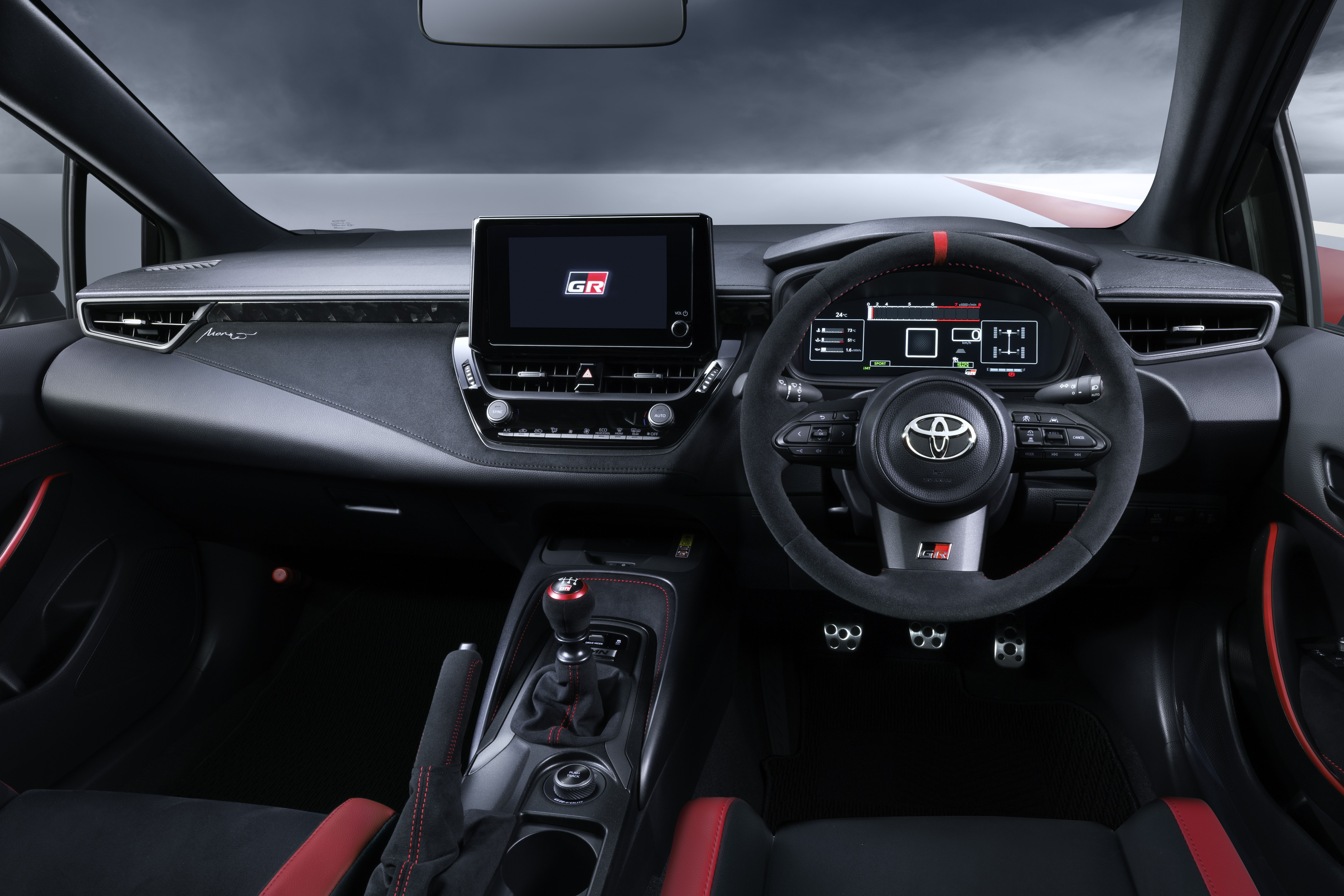 Interior de um carro esportivo com volante de camur&ccedil;a preta e faixa vermelha, painel digital, tela multim&iacute;dia com logo GR, c&acirc;mbio manual e bancos pretos com detalhes vermelhos