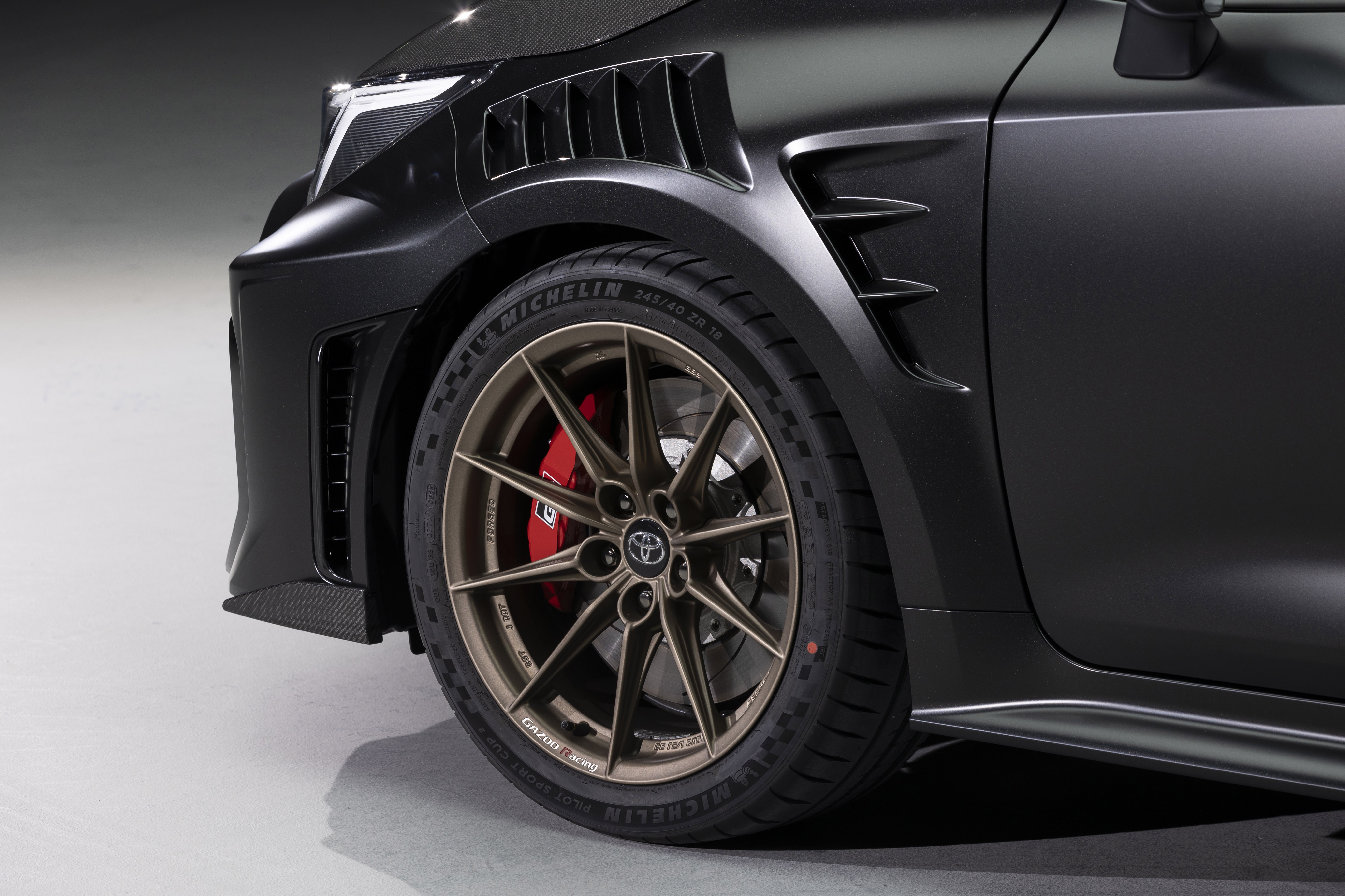 Lateral dianteira de um carro esportivo preto fosco, com farol LED branco aceso, grade frontal e sa&iacute;das de ar no para-lama. A roda de liga leve bronze tem pin&ccedil;as de freio vermelhas e pneus Michelin