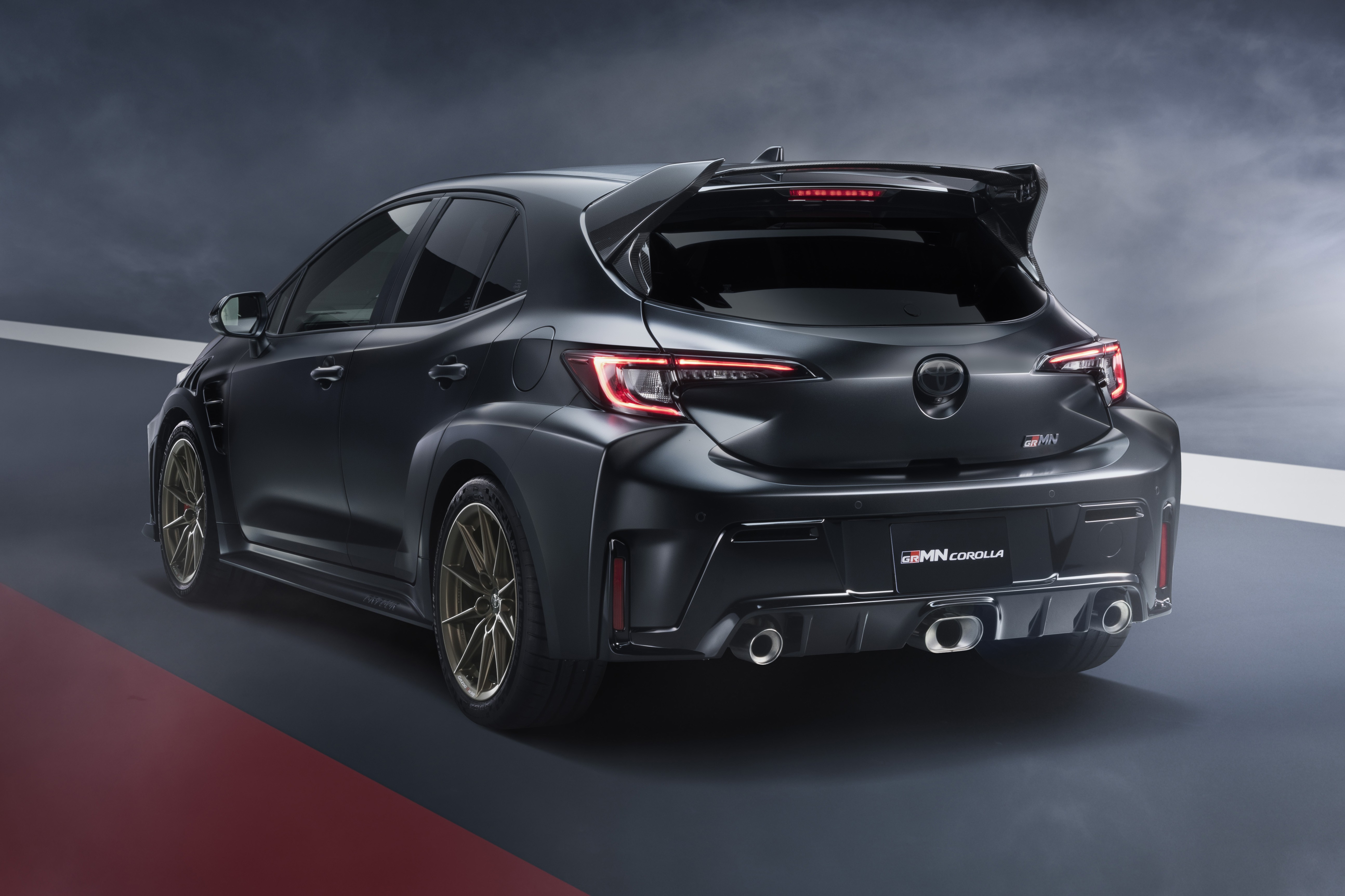 Toyota GRMN Corolla cinza escuro, visto de traseira e lateral, com rodas douradas, aerof&oacute;lio e tr&ecirc;s sa&iacute;das de escapamento, em uma pista escura com faixas vermelha e branca