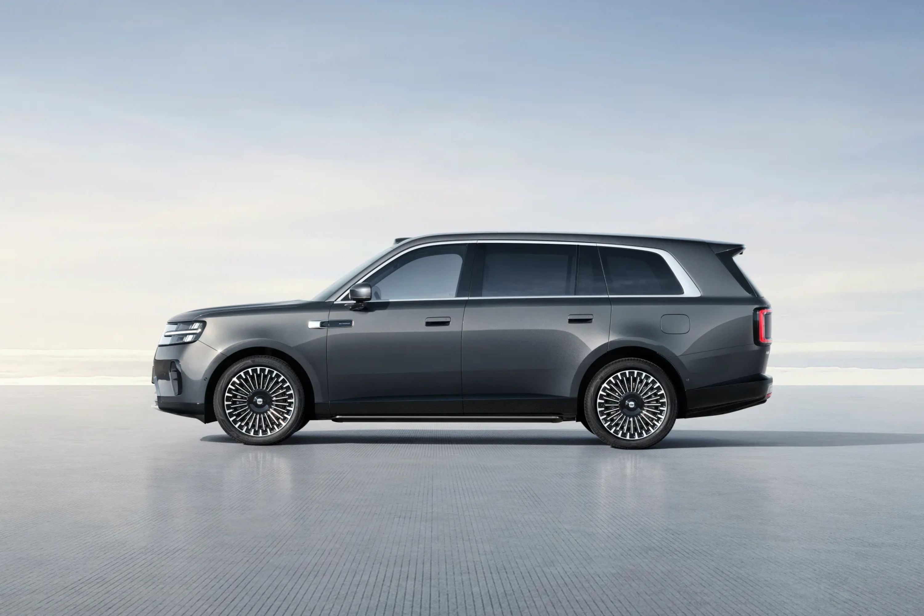 SUV cinza escuro, com rodas de design complexo em preto e prata, parado em uma superf&iacute;cie cinza clara sob um c&eacute;u azul p&aacute;lido
