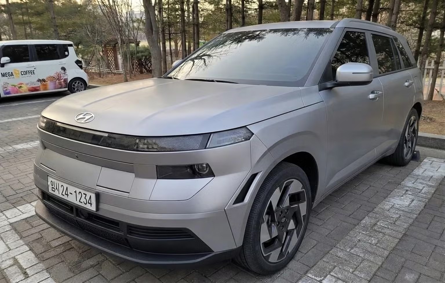 Carro Hyundai Kona cinza-prateado estacionado em piso de blocos de concreto, com uma van branca Mega Coffee ao fundo e &aacute;rvores sem folhas