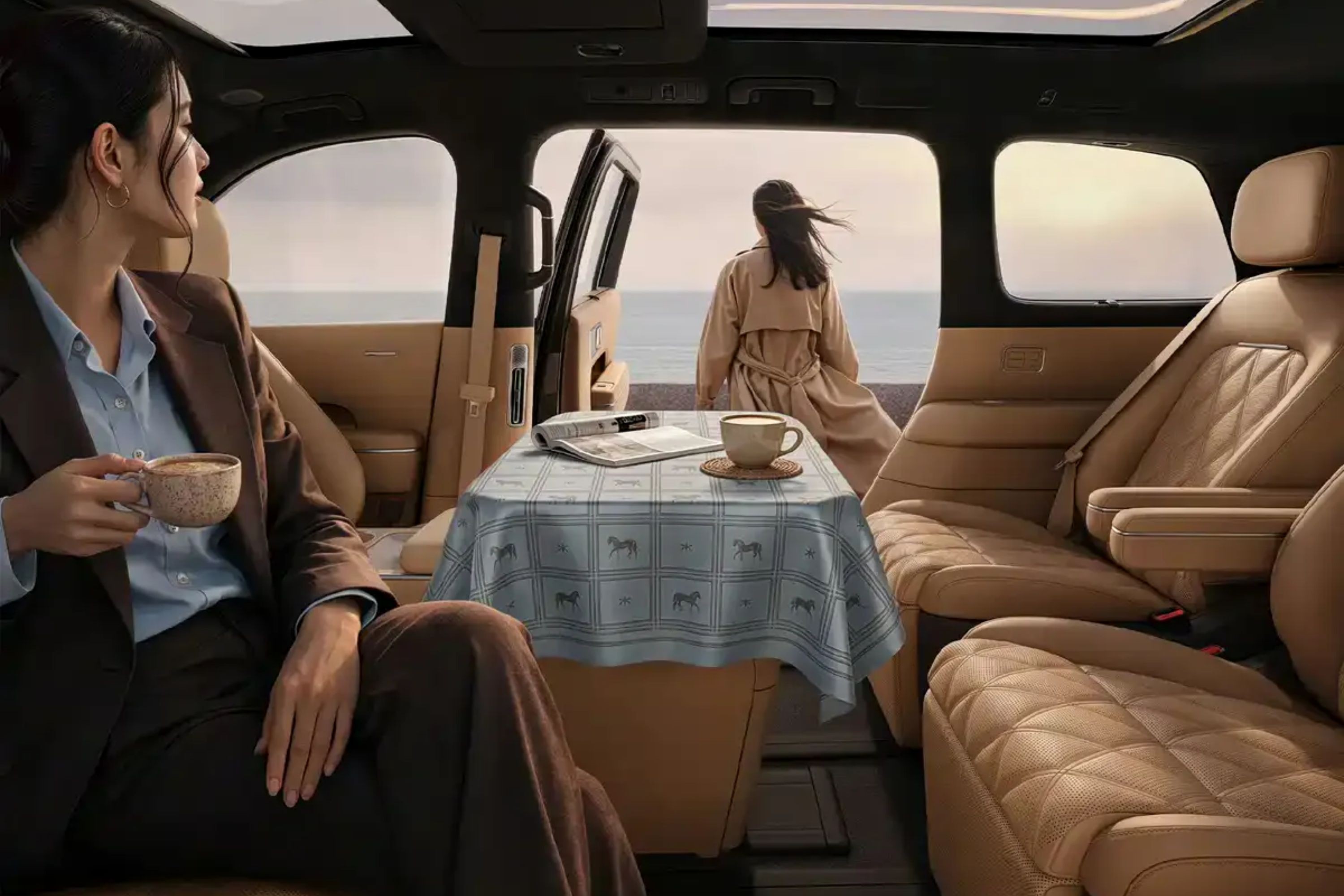 Mulher de terno marrom e camisa azul sentada em carro de luxo, segurando x&iacute;cara de caf&eacute;, olhando para outra mulher de casaco bege em frente ao mar. Uma mesa com toalha xadrez e x&iacute;cara de caf&eacute; est&aacute; entre elas