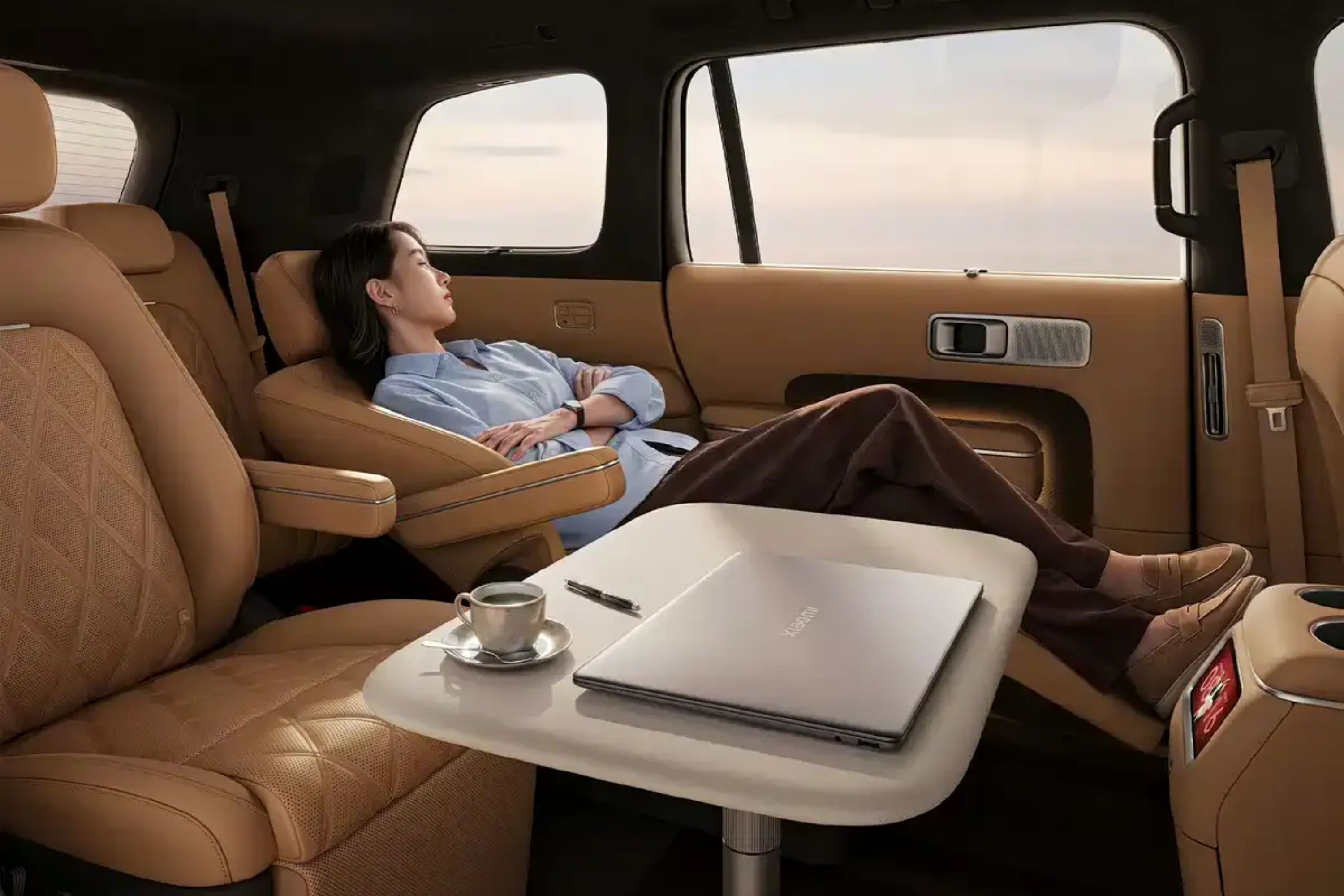 Mulher asi&aacute;tica de camisa azul e cal&ccedil;a marrom relaxa em assento reclin&aacute;vel de carro luxuoso, com os olhos fechados. Uma mesa lateral com notebook prateado, caneta e x&iacute;cara de caf&eacute; est&aacute; &agrave; sua frente. O interior do ve&iacute;culo &eacute; bege e marrom, com janelas mostrando um c&eacute;u claro ao fundo