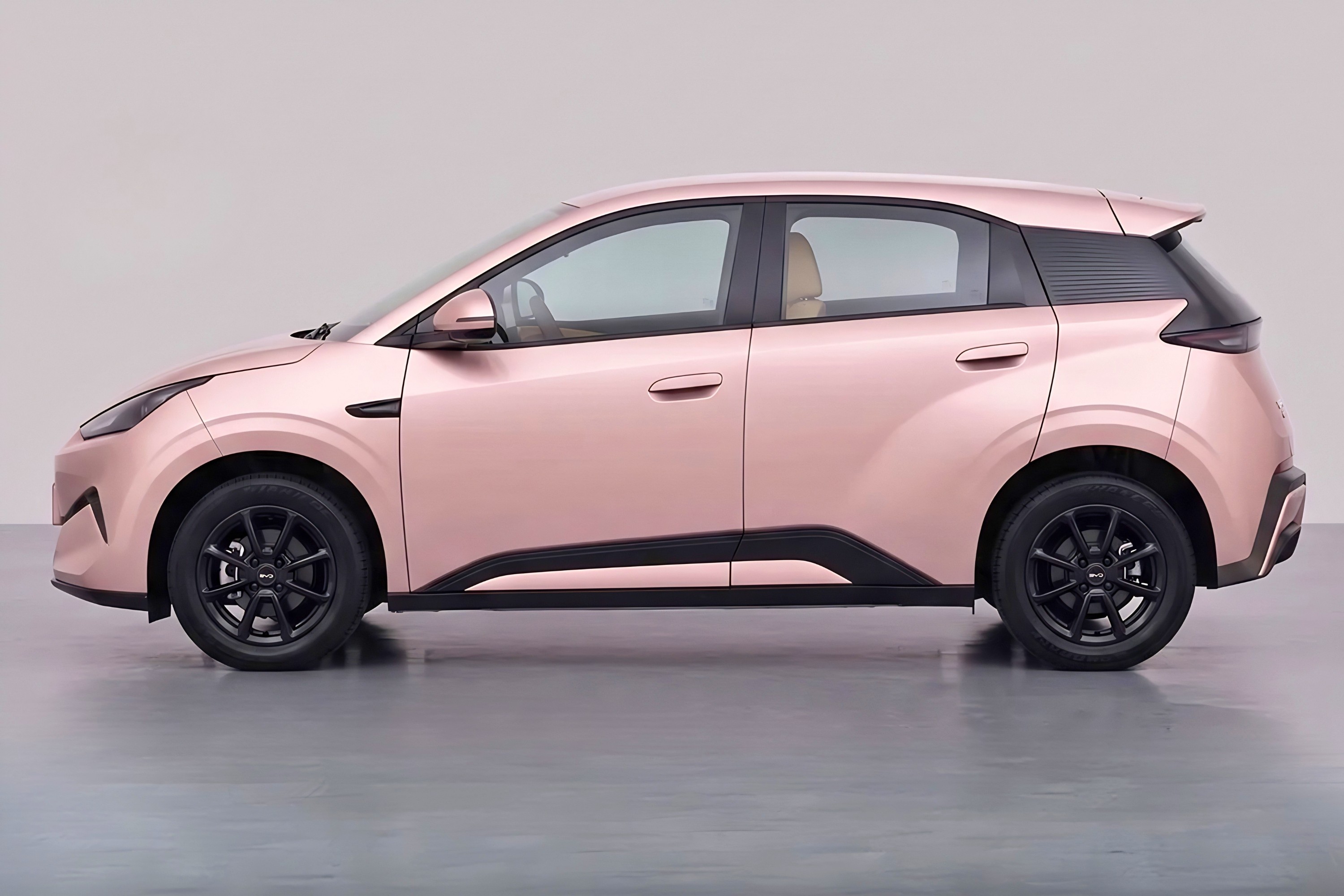 Carro compacto rosa met&aacute;lico, visto de perfil, com detalhes em preto fosco na parte inferior da carroceria e nas rodas. O interior claro &eacute; vis&iacute;vel pelas janelas. O ve&iacute;culo est&aacute; parado em um est&uacute;dio com fundo cinza claro e ch&atilde;o escuro.