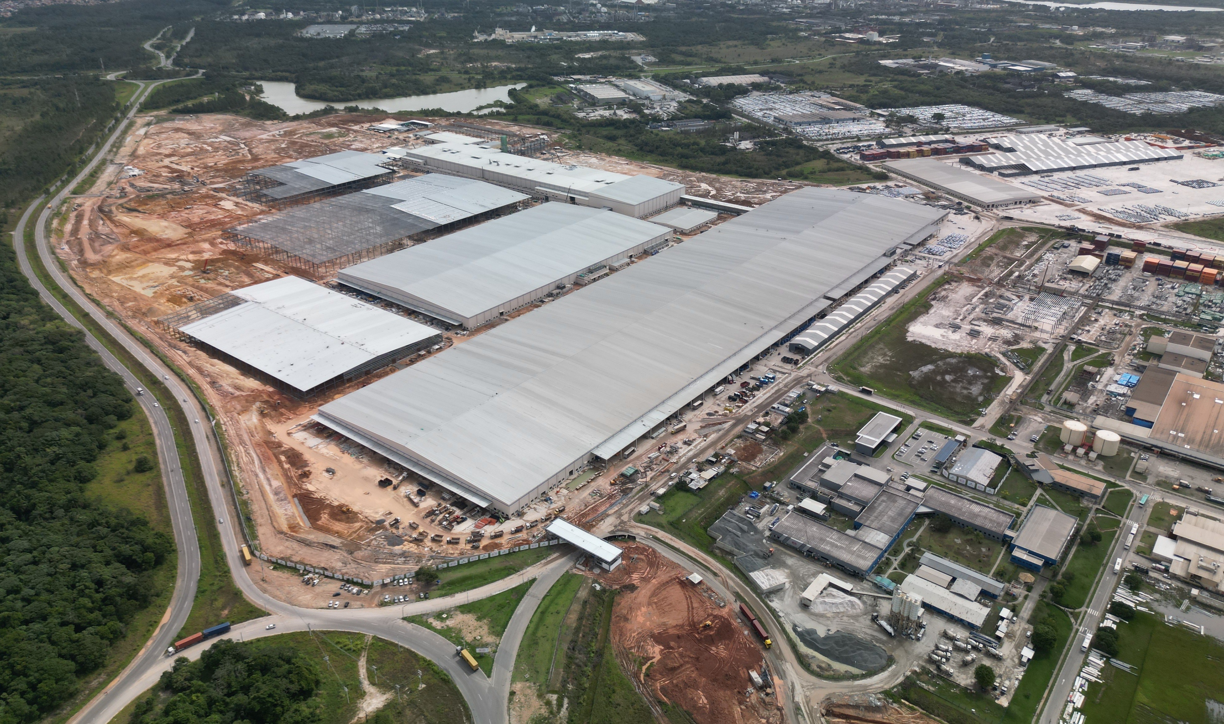 Vista a&eacute;rea de um grande complexo industrial em constru&ccedil;&atilde;o, com v&aacute;rios galp&otilde;es de telhado cinza, alguns j&aacute; cobertos e outros com estrutura met&aacute;lica aparente. H&aacute; &aacute;reas de terraplanagem e estradas sinuosas com ve&iacute;culos, cercadas por vegeta&ccedil;&atilde;o densa e um corpo d&aacute;gua ao fundo. Outras edifica&ccedil;&otilde;es menores e estacionamentos completam a paisagem industrial.