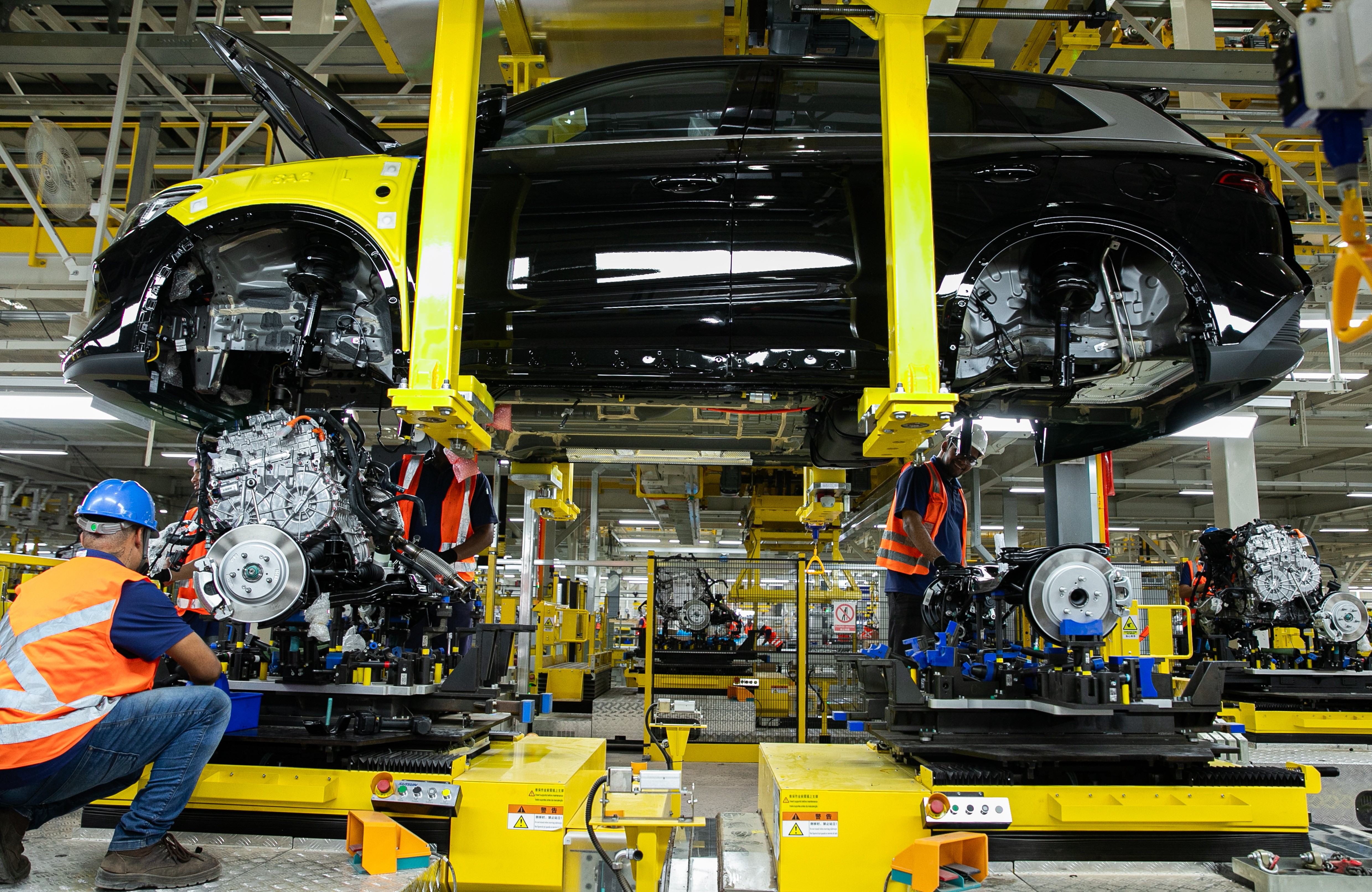 Homens em coletes laranja e capacetes azuis trabalham na montagem de carros em uma linha de produ&ccedil;&atilde;o industrial. Um carro preto suspenso por guindastes amarelos tem o cap&ocirc; aberto, enquanto os trabalhadores instalam motores e componentes em chassis. O ambiente &eacute; iluminado e repleto de equipamentos e m&aacute;quinas amarelas
