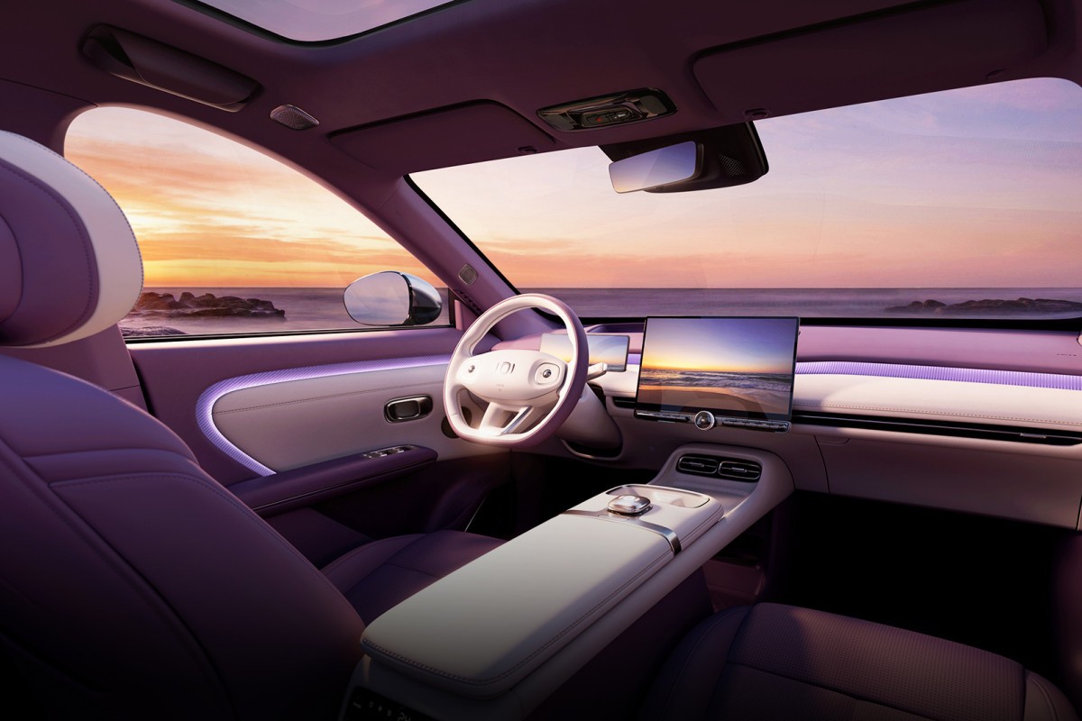 Interior de carro moderno com bancos roxos e detalhes brancos, volante branco e tela multim&iacute;dia exibindo p&ocirc;r do sol na praia. O painel tem ilumina&ccedil;&atilde;o ambiente roxa, e o teto solar panor&acirc;mico reflete o c&eacute;u alaranjado sobre o oceano com rochas &agrave; dist&acirc;ncia