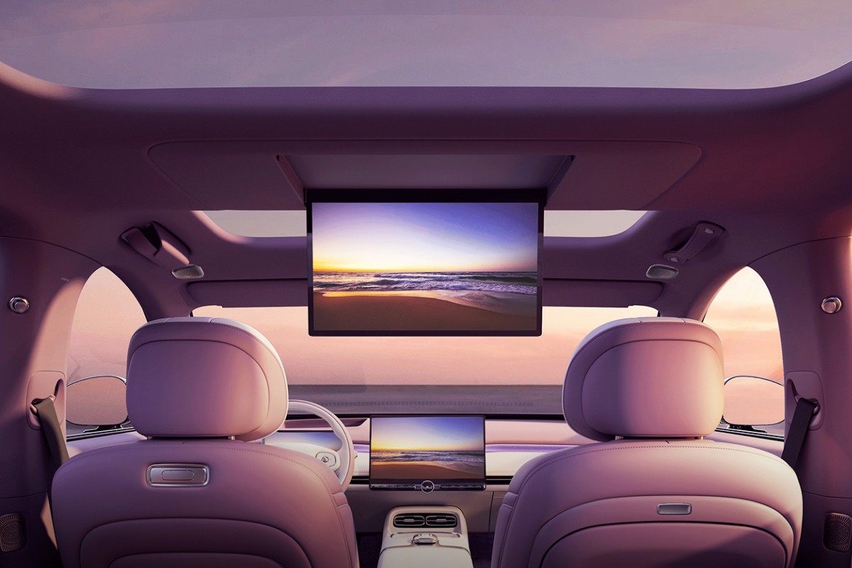 Interior de carro moderno com bancos roxos claros, teto solar e duas telas exibindo um p&ocirc;r do sol na praia. Uma tela grande desce do teto, e outra menor est&aacute; no painel central. O volante branco &eacute; vis&iacute;vel &agrave; esquerda, e o c&eacute;u rosado &eacute; visto pelas janelas