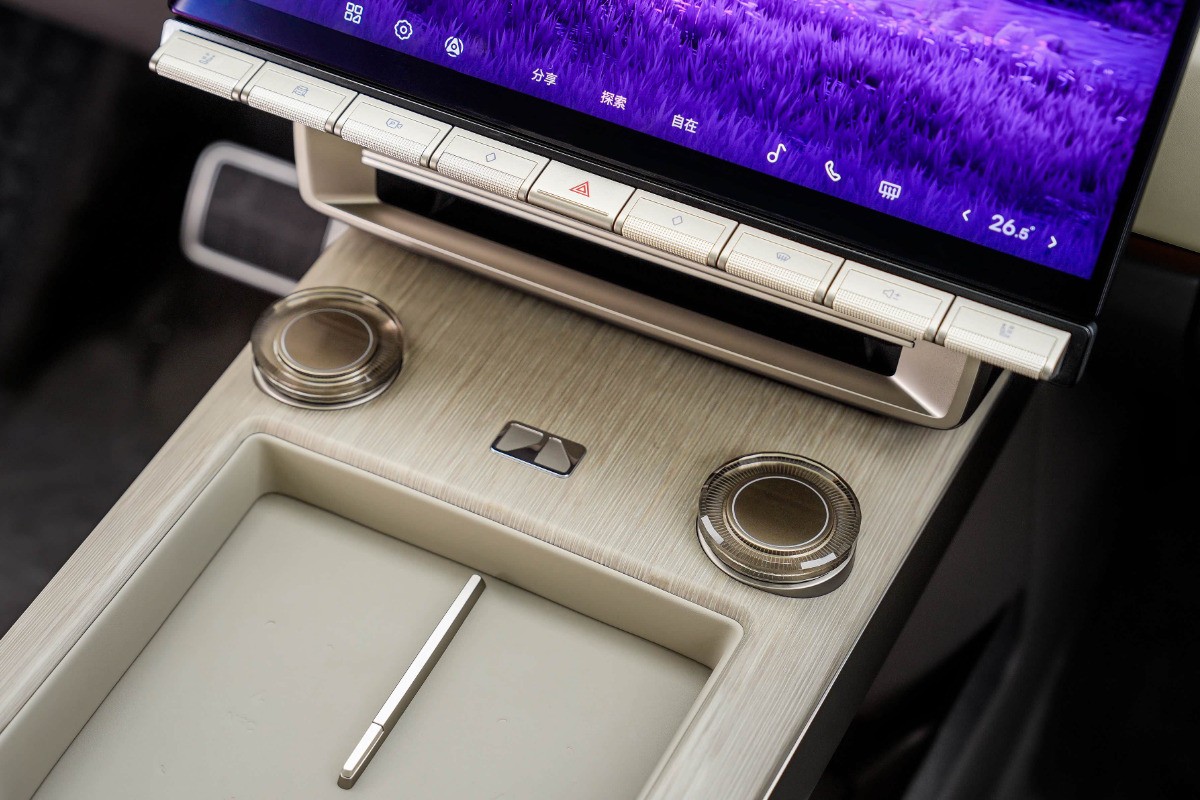 Painel de controle de carro com tela sens&iacute;vel ao toque exibindo um fundo roxo, bot&otilde;es f&iacute;sicos prateados e dois bot&otilde;es girat&oacute;rios transparentes. Uma caneta stylus prateada repousa em um compartimento abaixo