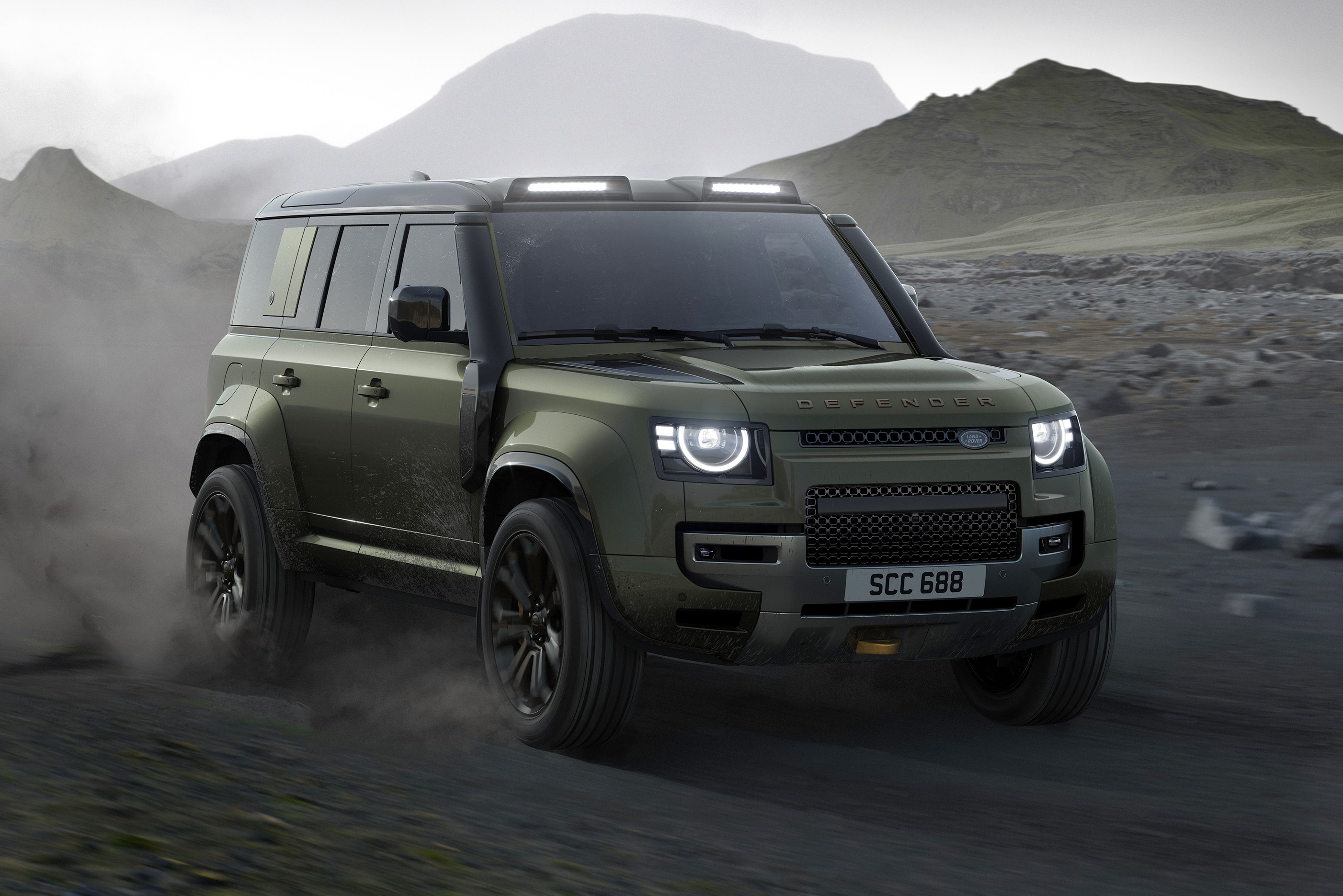 SUV Land Rover Defender verde-oliva em movimento, levantando poeira em uma estrada de terra. O ve&iacute;culo tem far&oacute;is acesos, luzes auxiliares no teto e snorkel lateral. Ao fundo, montanhas rochosas sob um c&eacute;u nublado