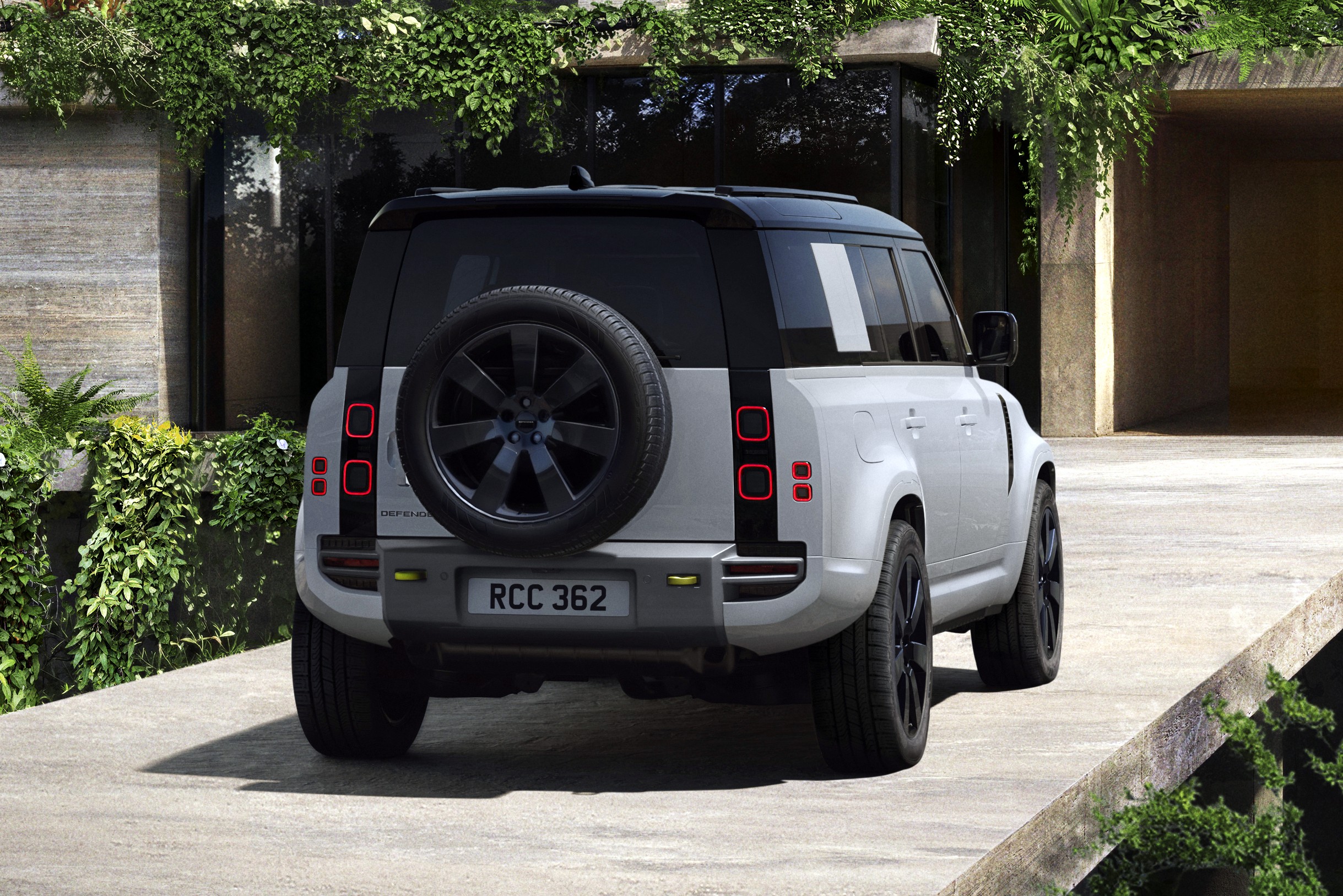 Traseira de um Land Rover Defender branco com teto preto, pneu sobressalente na porta traseira e lanternas quadradas vermelhas, estacionado em uma rampa de concreto com vegeta&ccedil;&atilde;o exuberante em um edif&iacute;cio moderno ao fundo