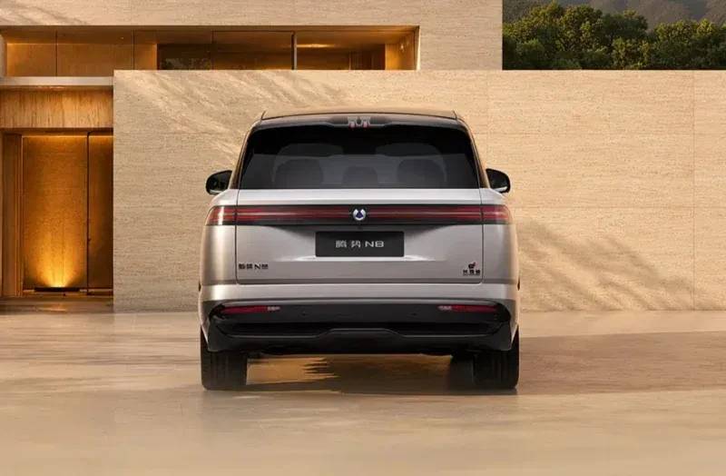 Traseira de um carro SUV cinza-prateado, com lanternas horizontais e um logotipo circular no centro, estacionado em frente a uma parede de pedra clara com uma entrada iluminada &agrave; esquerda e vegeta&ccedil;&atilde;o no topo &agrave; direita