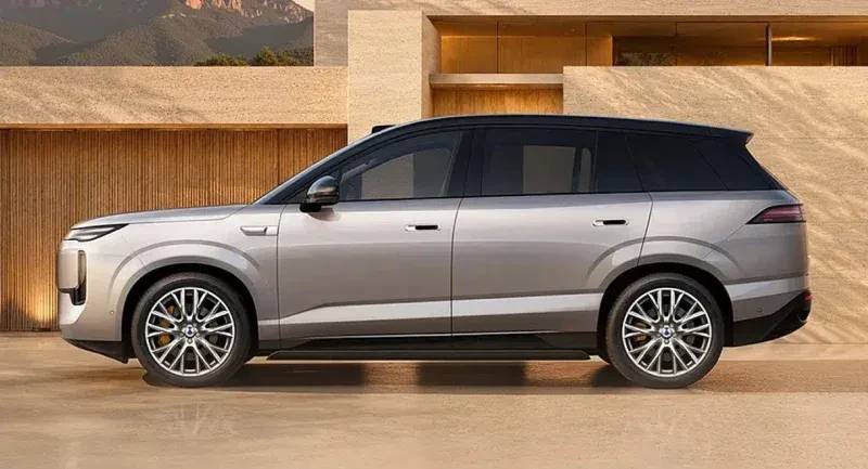 SUV cinza claro com teto preto, rodas esportivas prateadas e detalhes cromados, estacionado de perfil em frente a uma casa moderna com paredes de madeira e pedra, e montanhas ao fundo