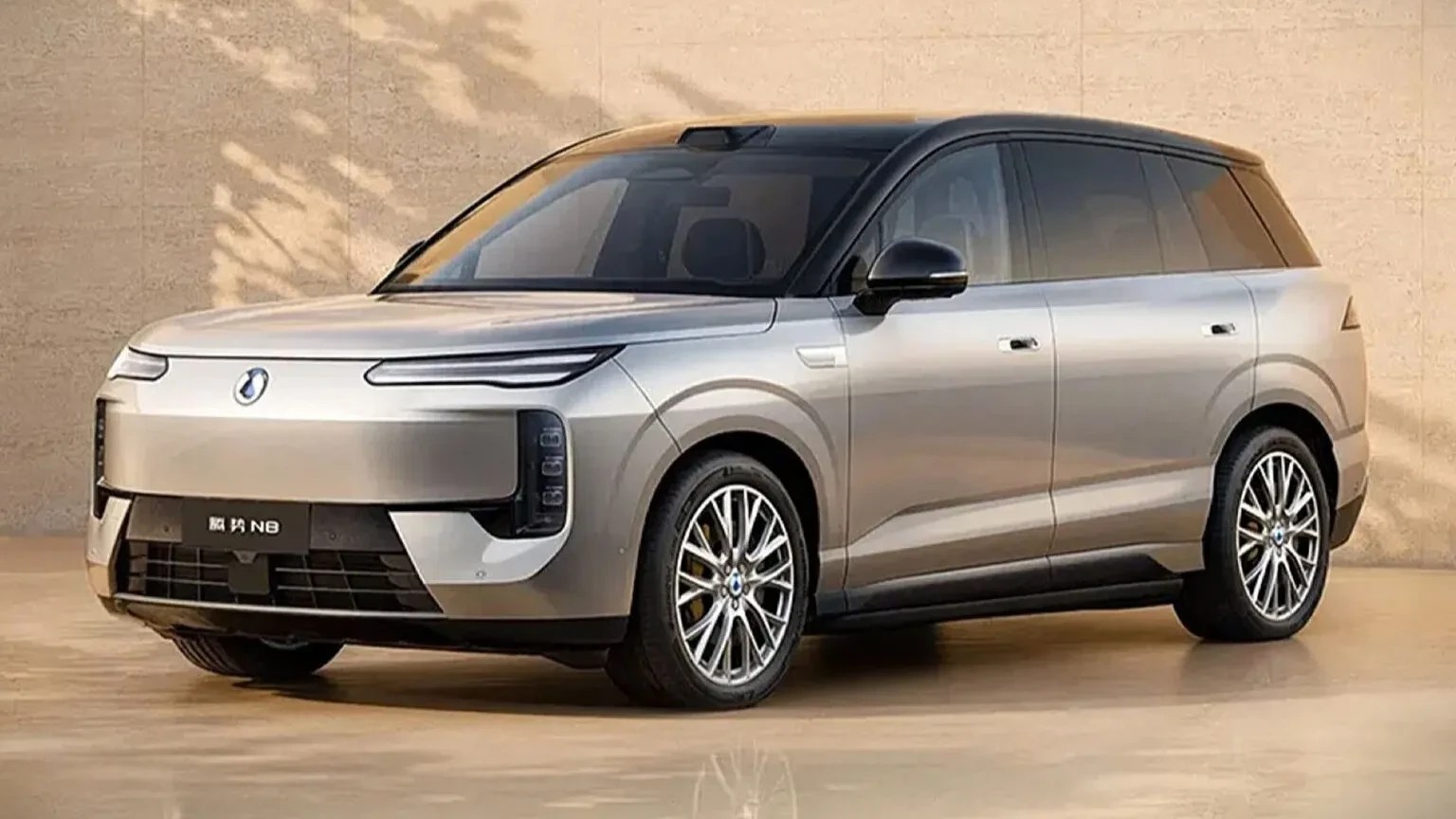 SUV cinza claro com teto preto, estacionado em um piso bege. O carro tem faróis finos e uma grade frontal fechada, com o emblema da marca no centro. As rodas são prateadas com múltiplos raios. A lateral do veículo exibe maçanetas discretas e janelas escurecidas. Ao fundo, uma parede bege com sombras de folhagens.