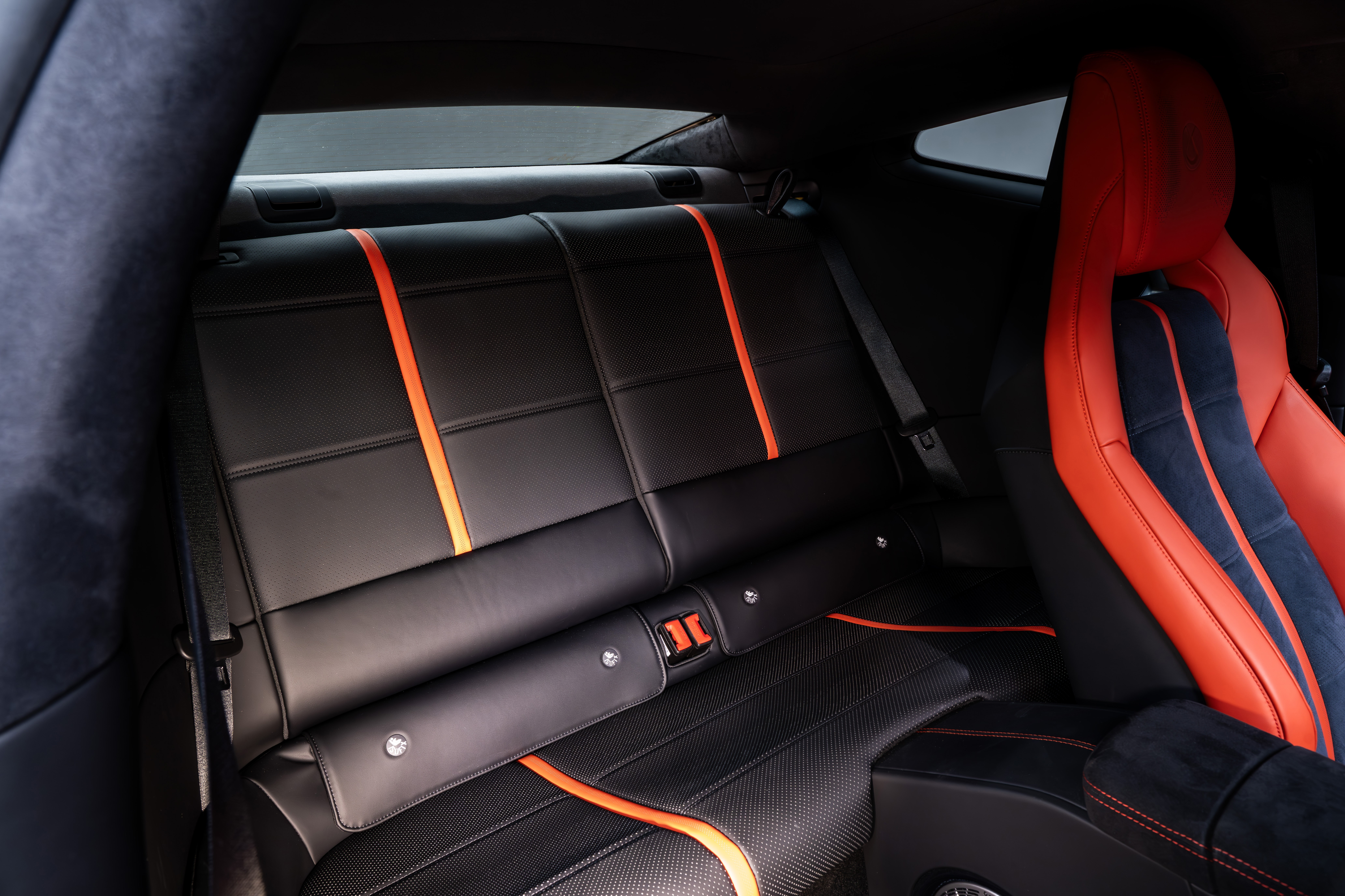 Interior de um carro esportivo com bancos traseiros pretos de couro, detalhados com faixas verticais e horizontais laranja. O banco dianteiro direito, vis&iacute;vel parcialmente, &eacute; vermelho com detalhes em azul escuro. O cinto de seguran&ccedil;a traseiro est&aacute; vis&iacute;vel, e o ambiente &eacute; escuro, com luz vindo das janelas