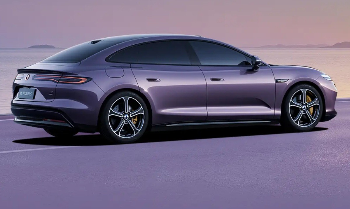 Carro el&eacute;trico roxo escuro, modelo sedan, estacionado em uma estrada roxa com o mar e montanhas ao fundo, sob um c&eacute;u alaranjado ao p&ocirc;r do sol