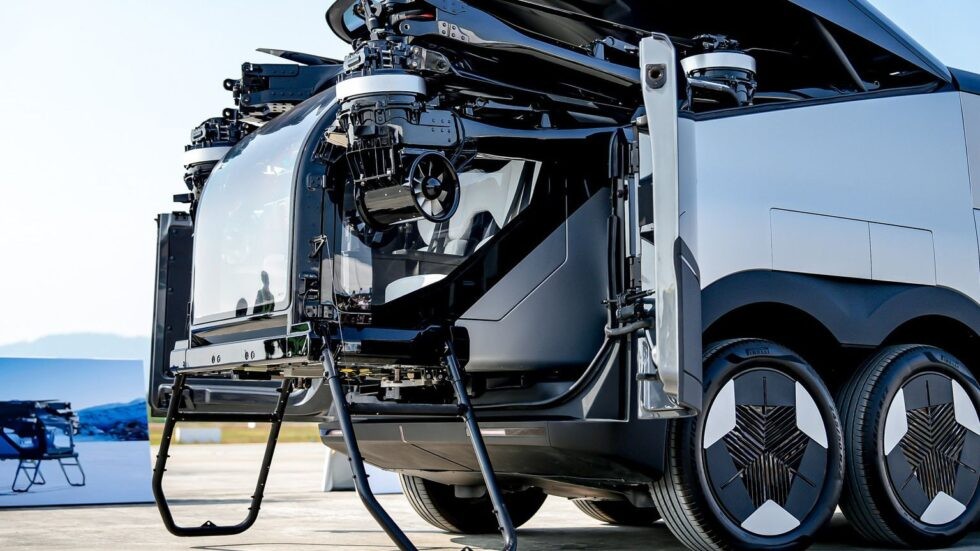 Ve&iacute;culo futurista preto e prata, com cabine de vidro e h&eacute;lices vis&iacute;veis na parte superior, em um ambiente externo com c&eacute;u claro. O ve&iacute;culo possui rodas com design inovador, e uma estrutura de pouso preta se estende da cabine. Ao fundo, uma imagem de montanhas e um lago.
