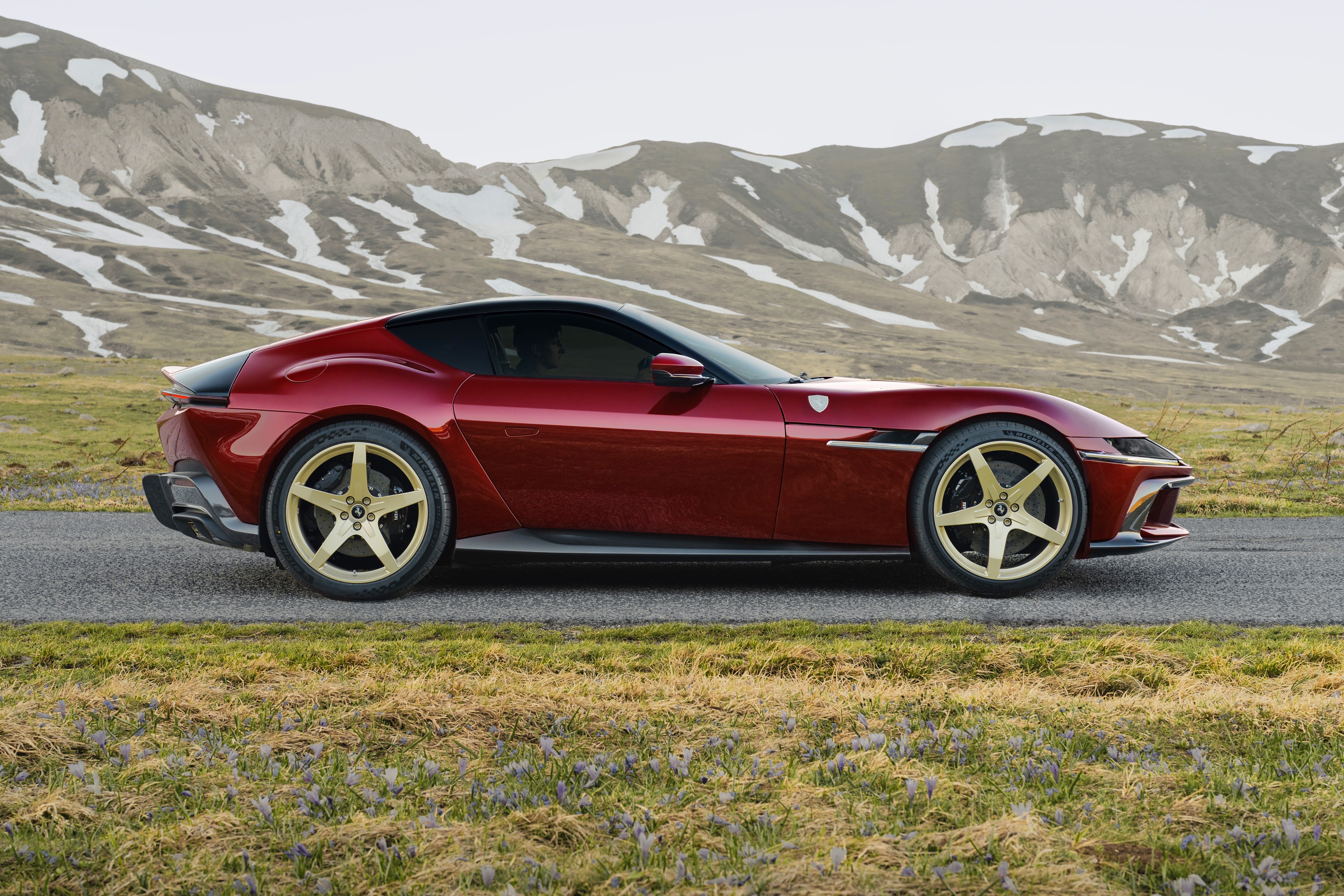 Carro esportivo vermelho escuro com teto preto e rodas douradas de cinco raios, estacionado em uma estrada asfaltada. Ao fundo, montanhas com picos nevados e vegeta&ccedil;&atilde;o rasteira com flores roxas no primeiro plano