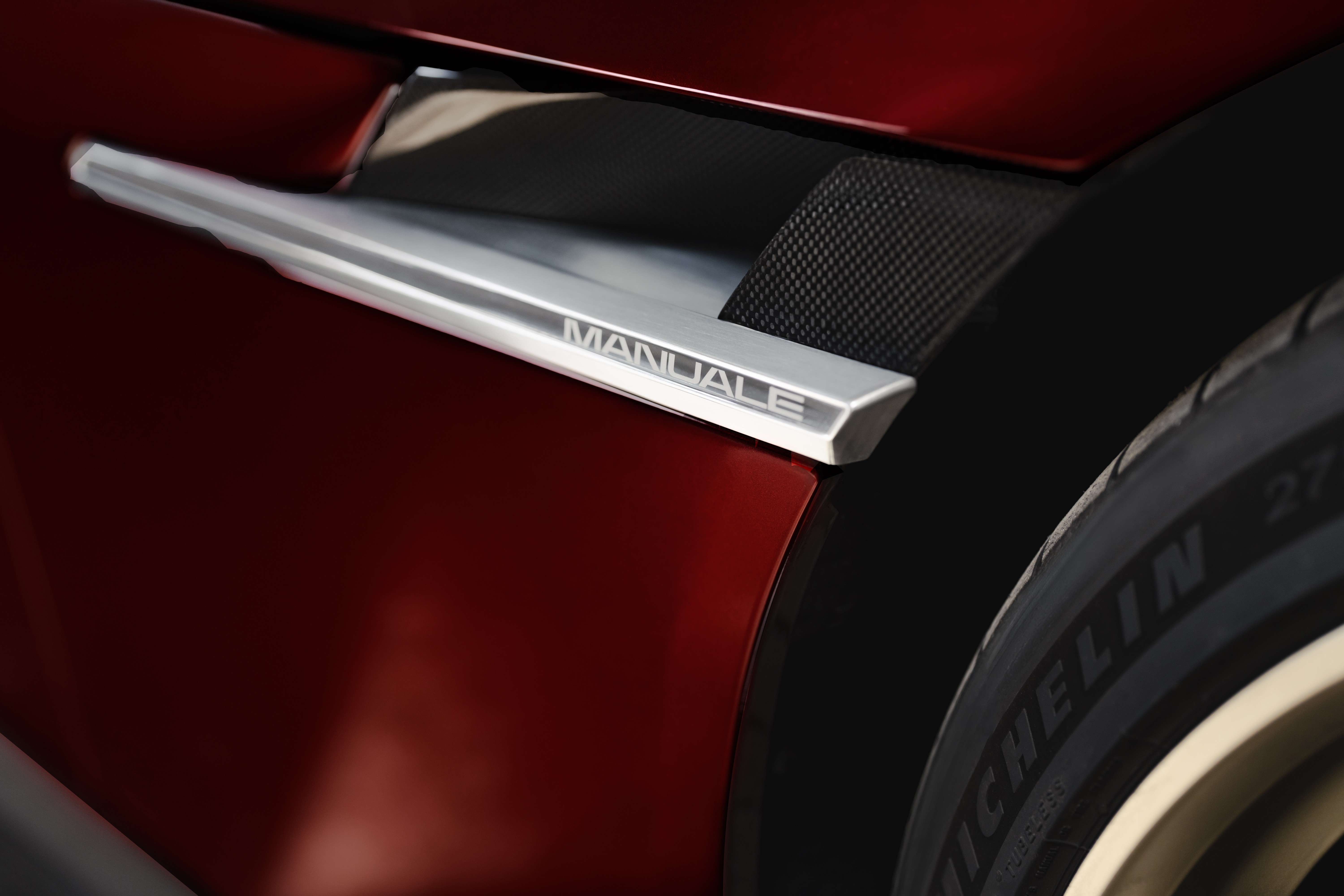 Detalhe da lateral de um carro esportivo vermelho-escuro, com um emblema prateado escrito MANUALE e uma grade preta texturizada. Parte de um pneu Michelin &eacute; vis&iacute;vel &agrave; direita
